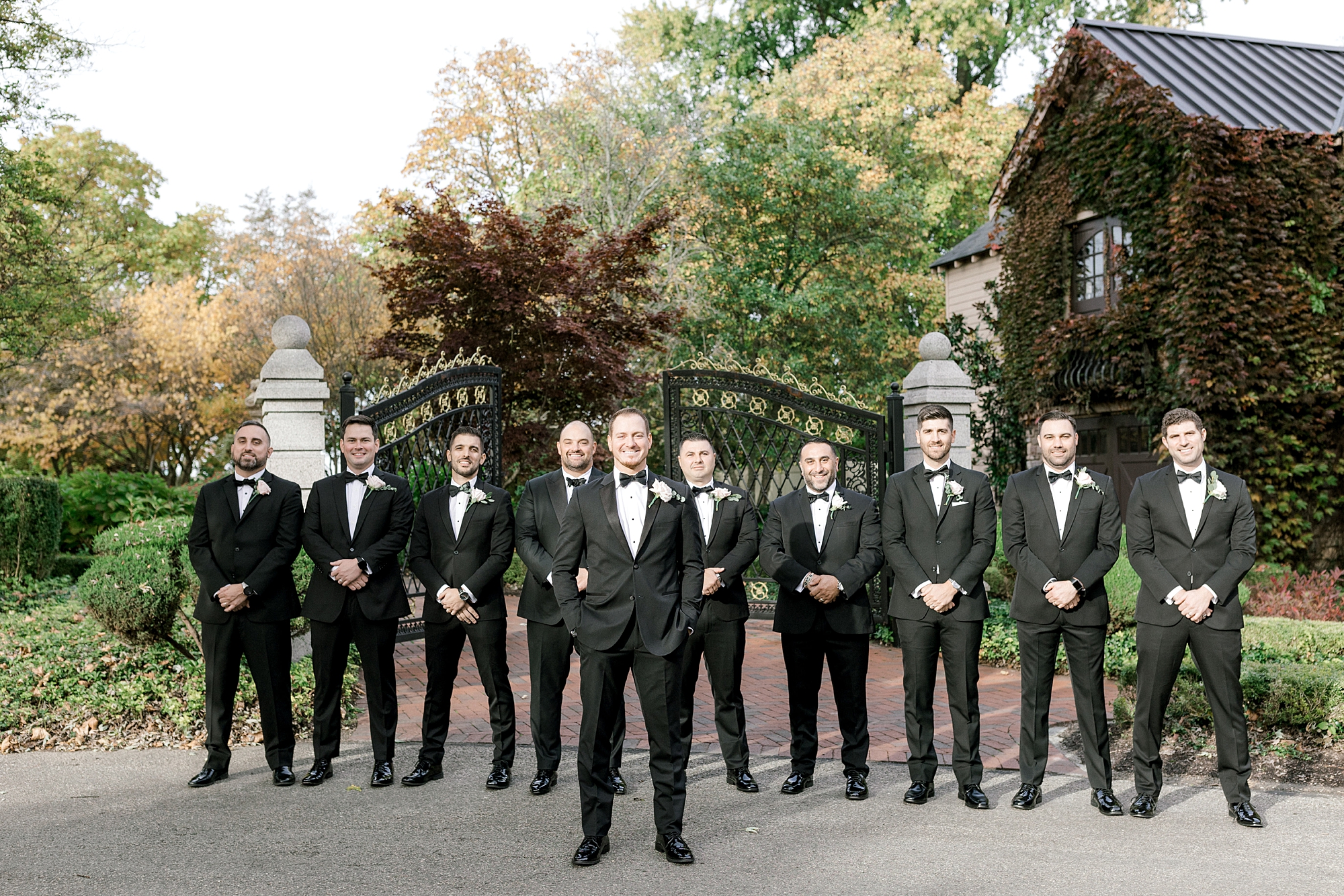 groom stands in front of groomsmen in black suits by metal gate at the Ashford Estate