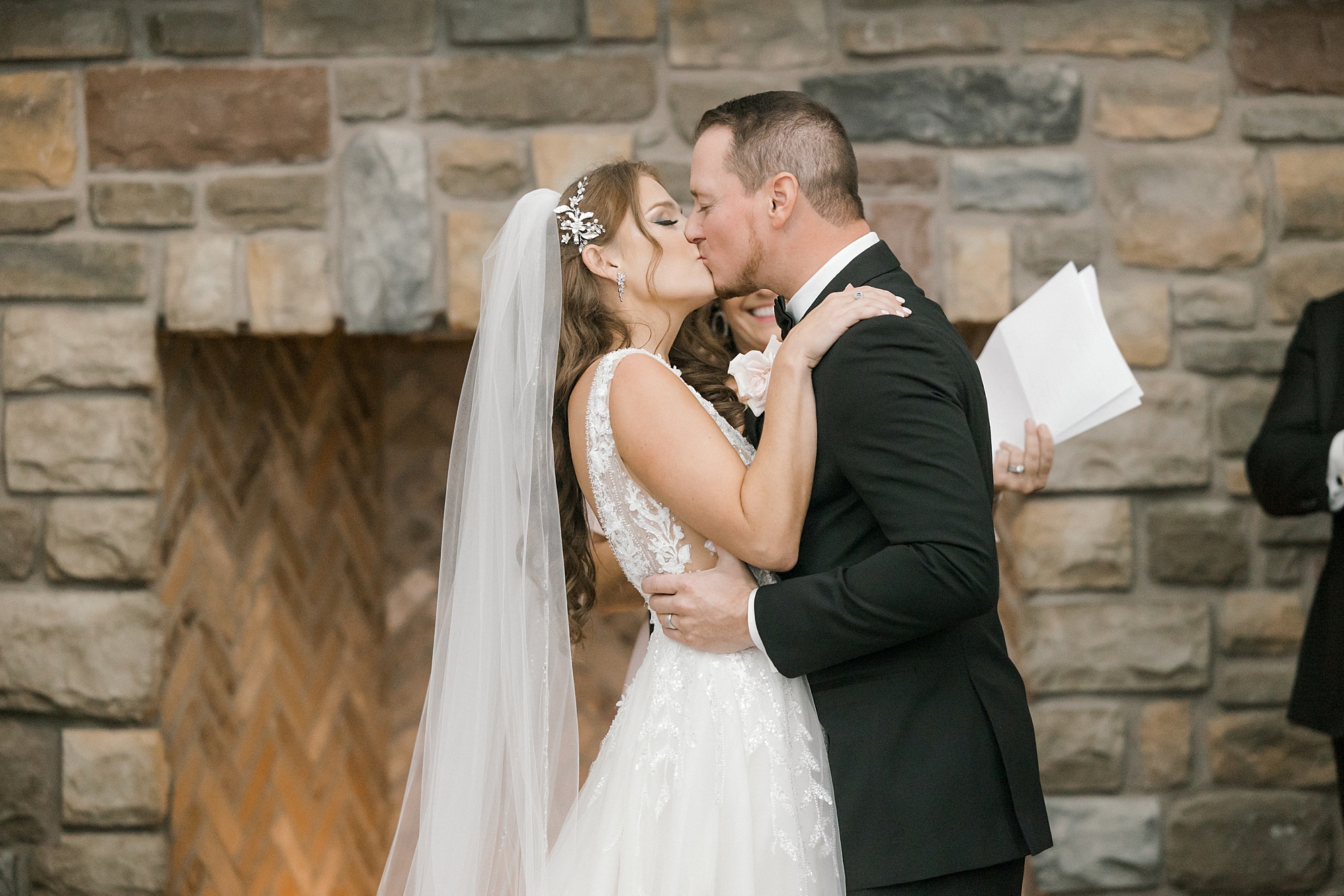 bride and groom kiss during wedding ceremony by stone fireplace