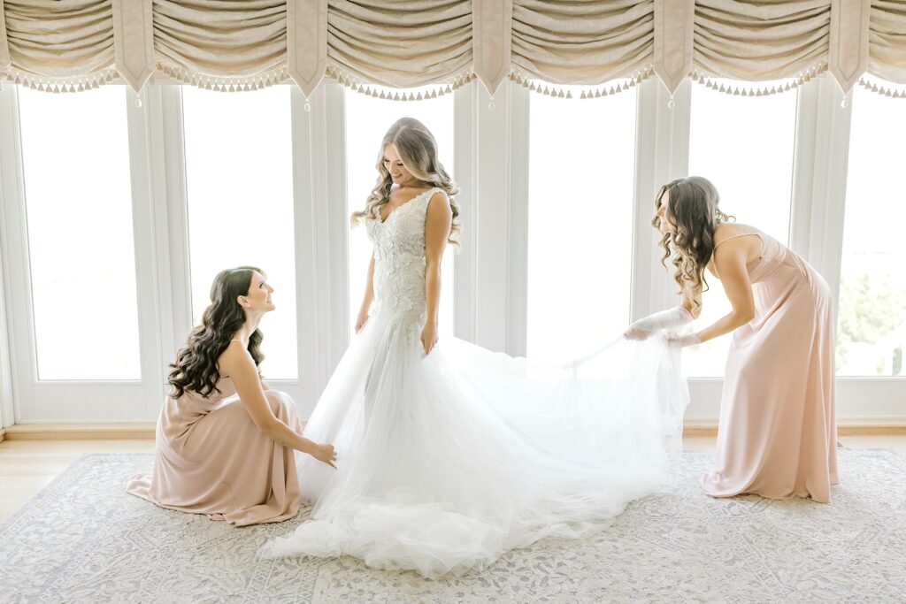 bridesmaids in dusty pink dresses adjusts bride's skirt by window 