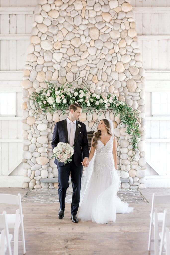 bride and groom hold hands walking by stone fireplace 