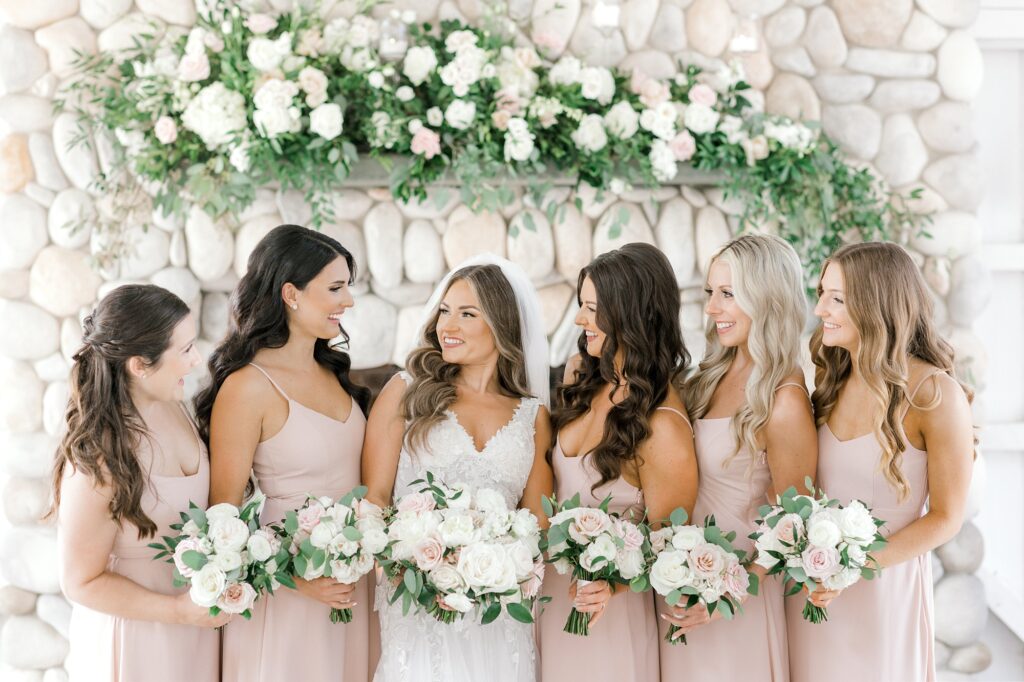 bride and bridesmaids in pink gowns smile in front of ivory flowers on stone fireplace 