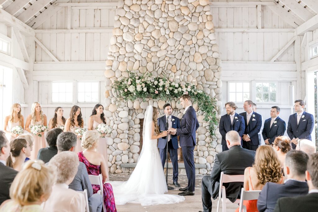 bride and groom hold hands by stone fireplace during ceremony at Bonnet Island Estate