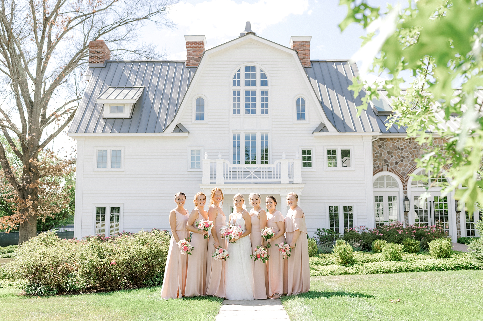 bride stands with bridesmaids in pink gowns by white house at Ryland Inn