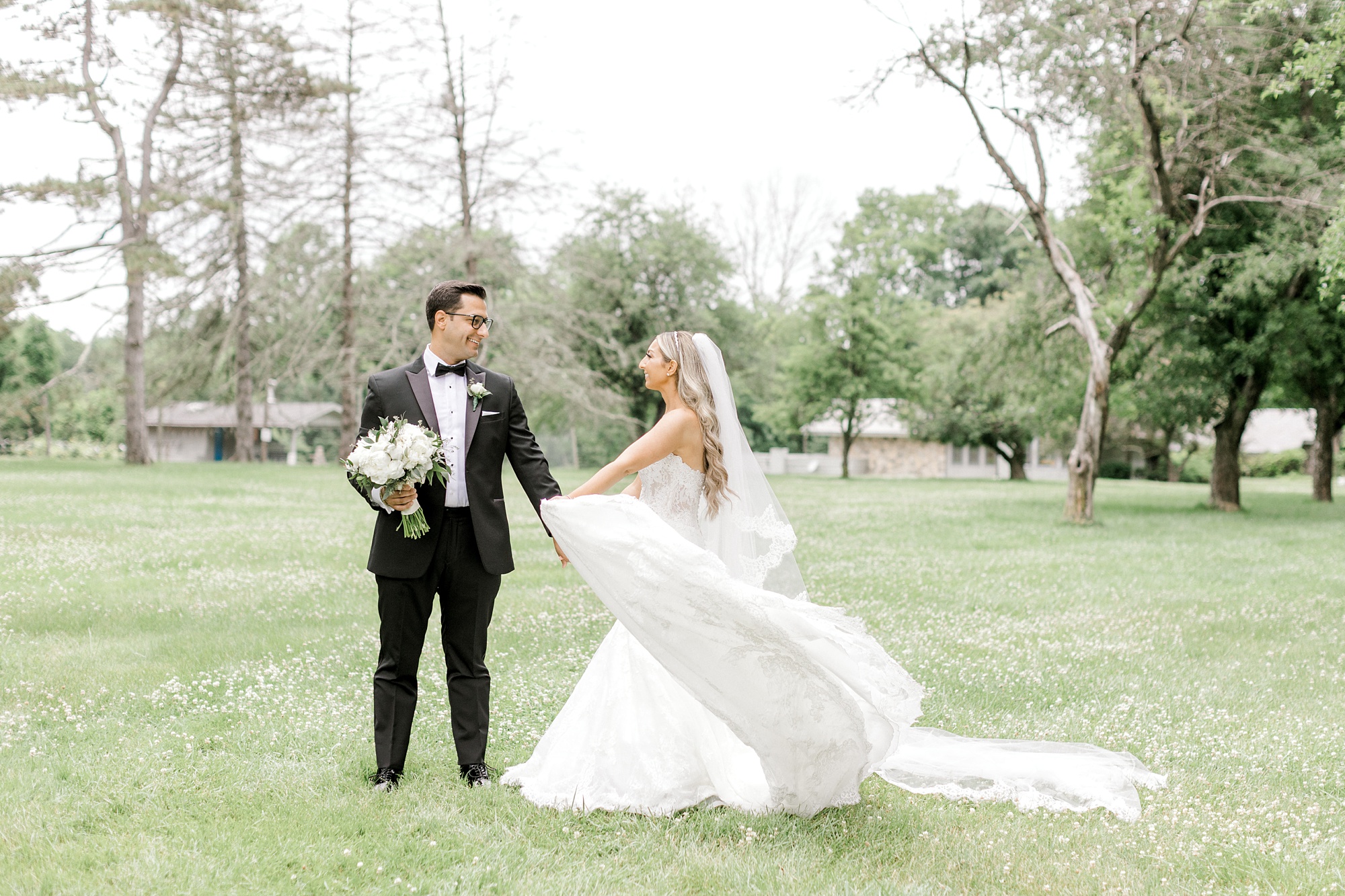 groom walks with bride on lawn while she twirls skirt of wedding gown