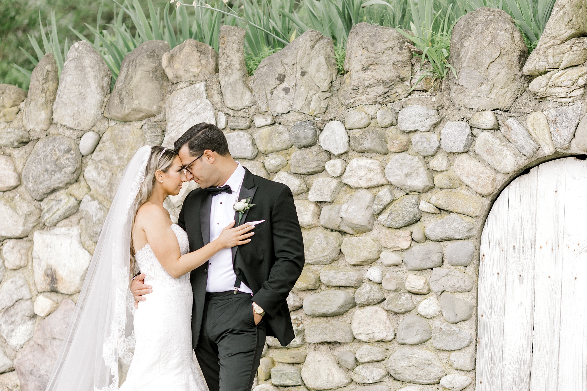 bride and groom lean heads together along stone wall at The Refinery at Perona Farms