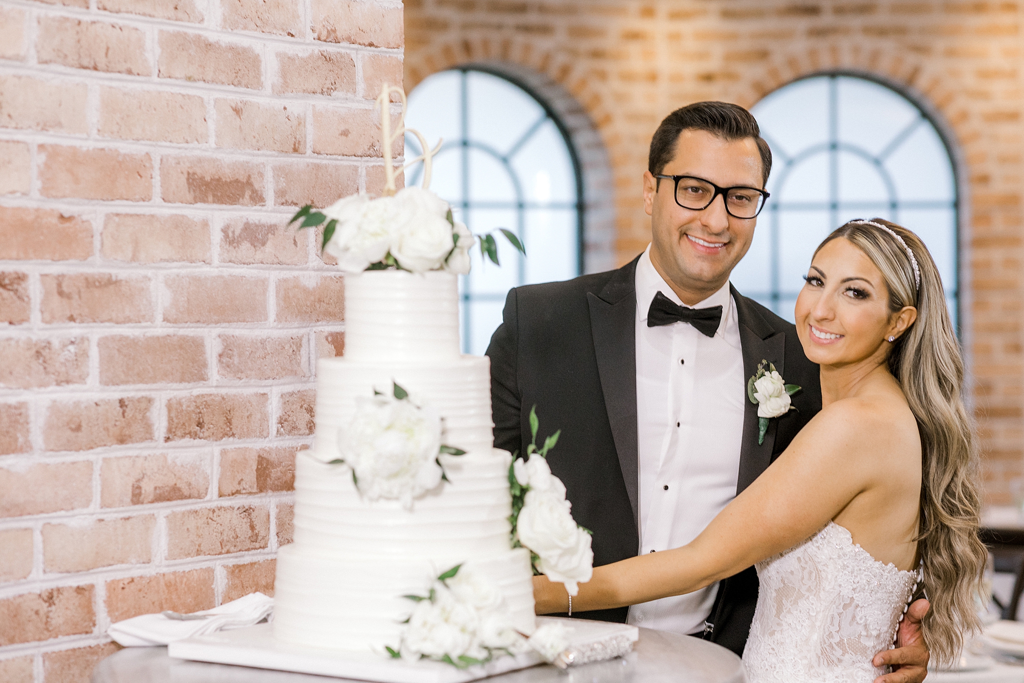 bride and groom pose by tiered wedding cake by brick wall