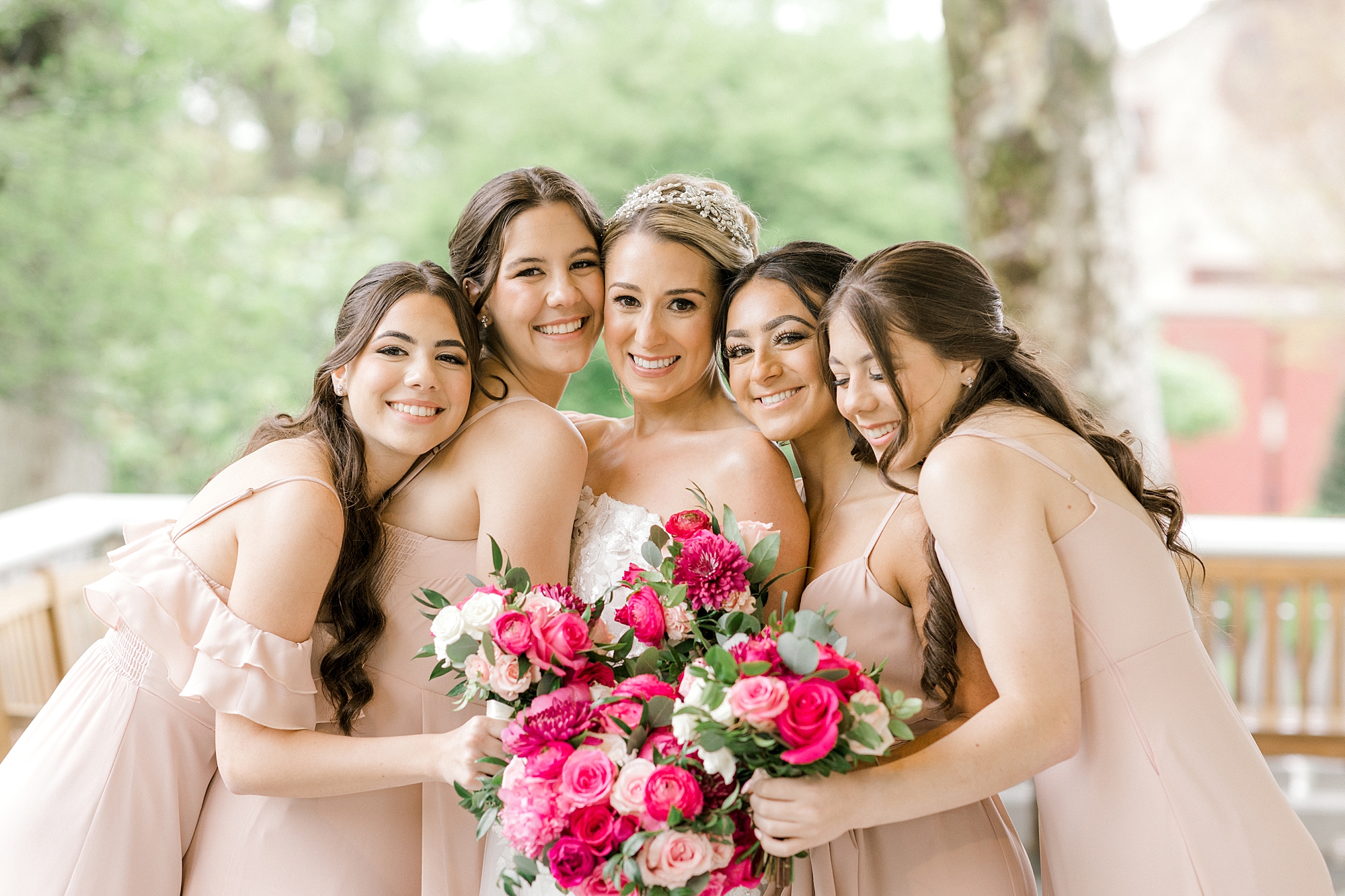 bridesmaids in blush gowns hug bride holding bouquets of bright pink roses