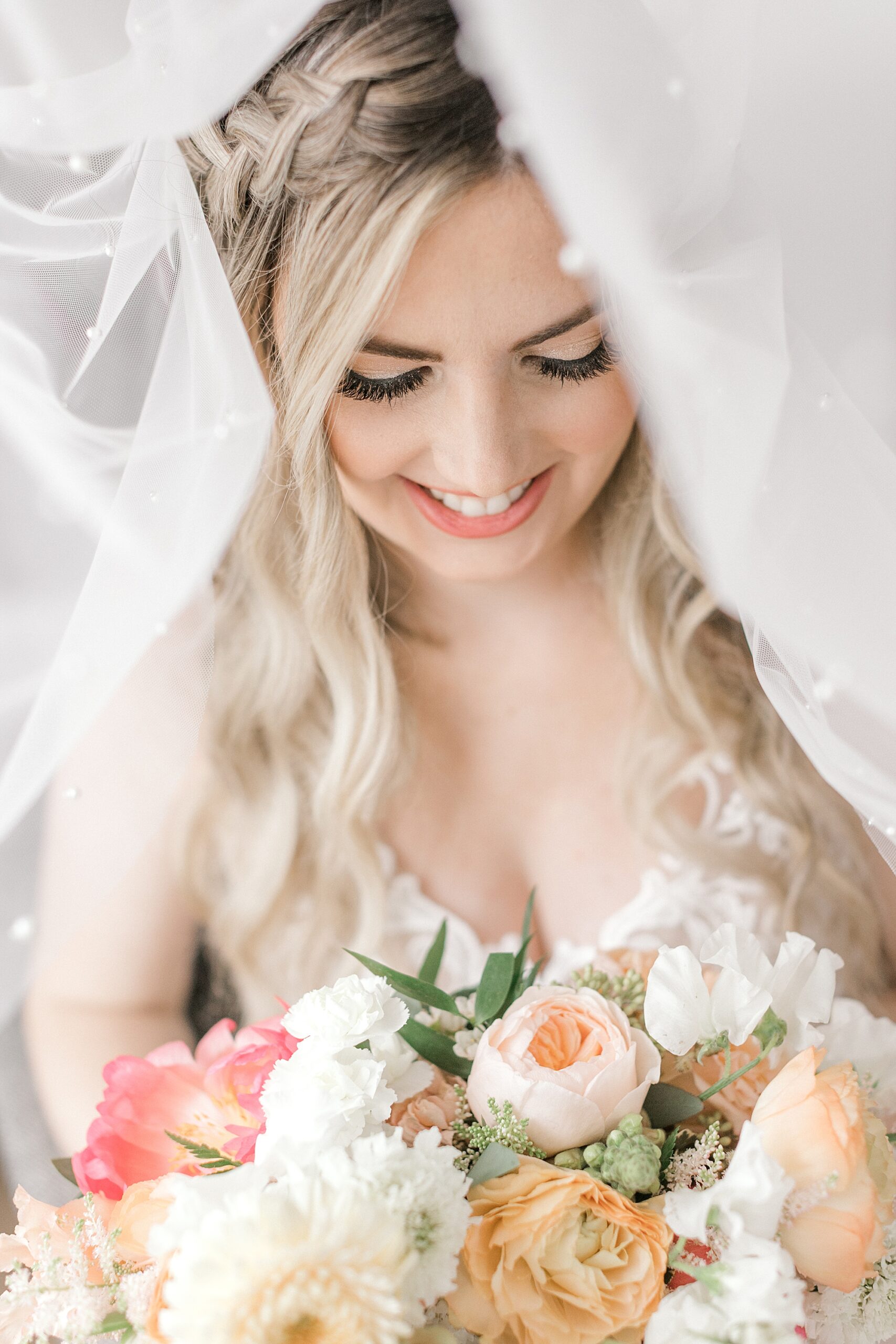blonde bride looks down at spring bouquet of ivory and peach flowers