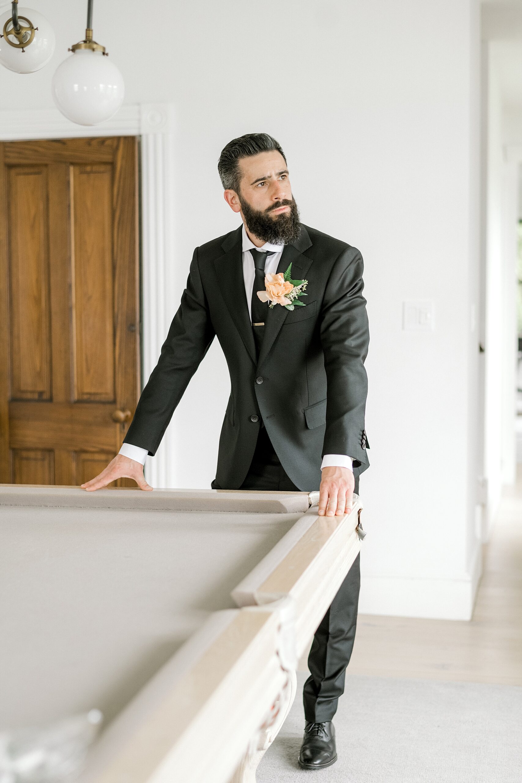 groom in black suit leans on edge of white pool table