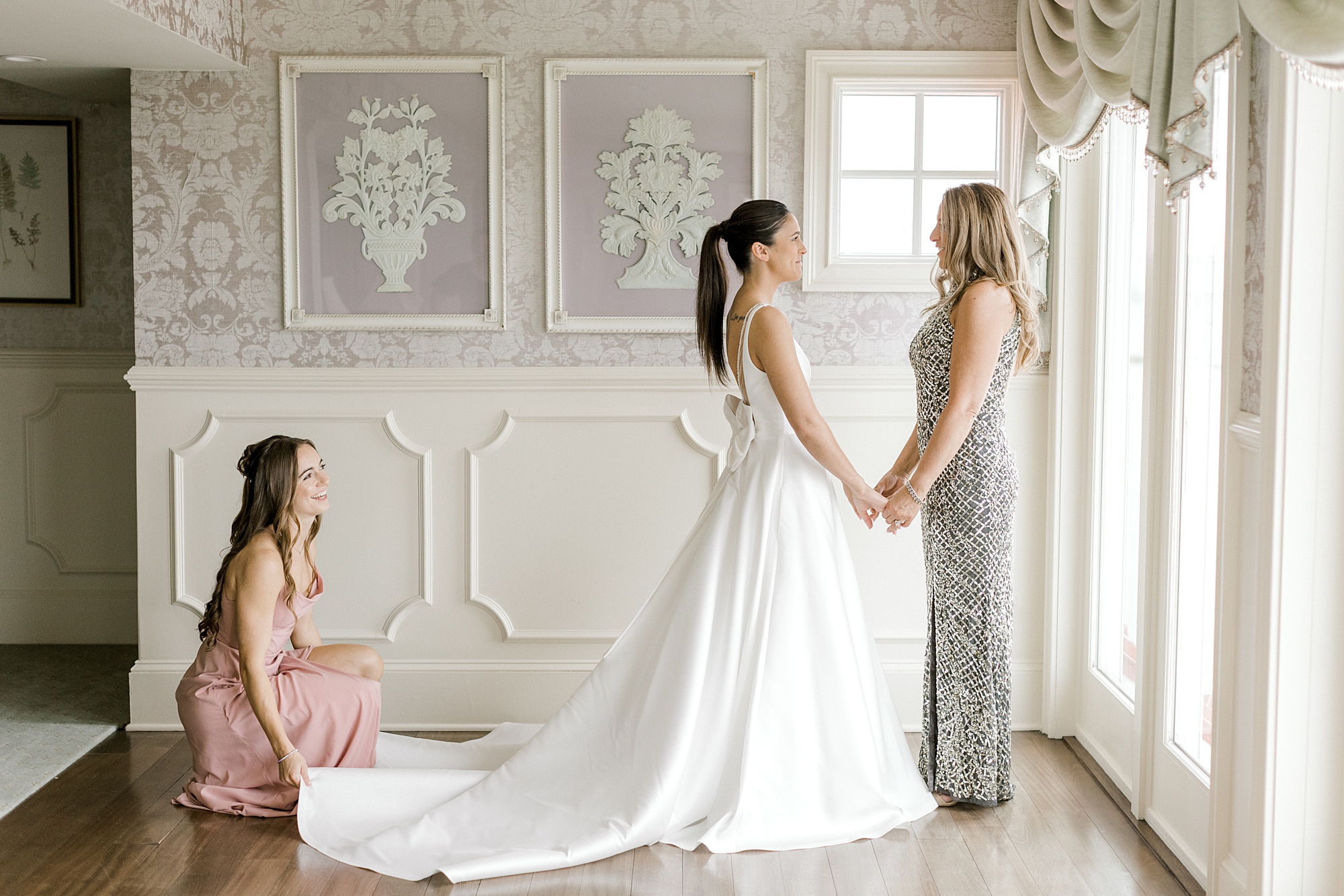 bridesmaid helps bride with skirt while bride holds hands with mom