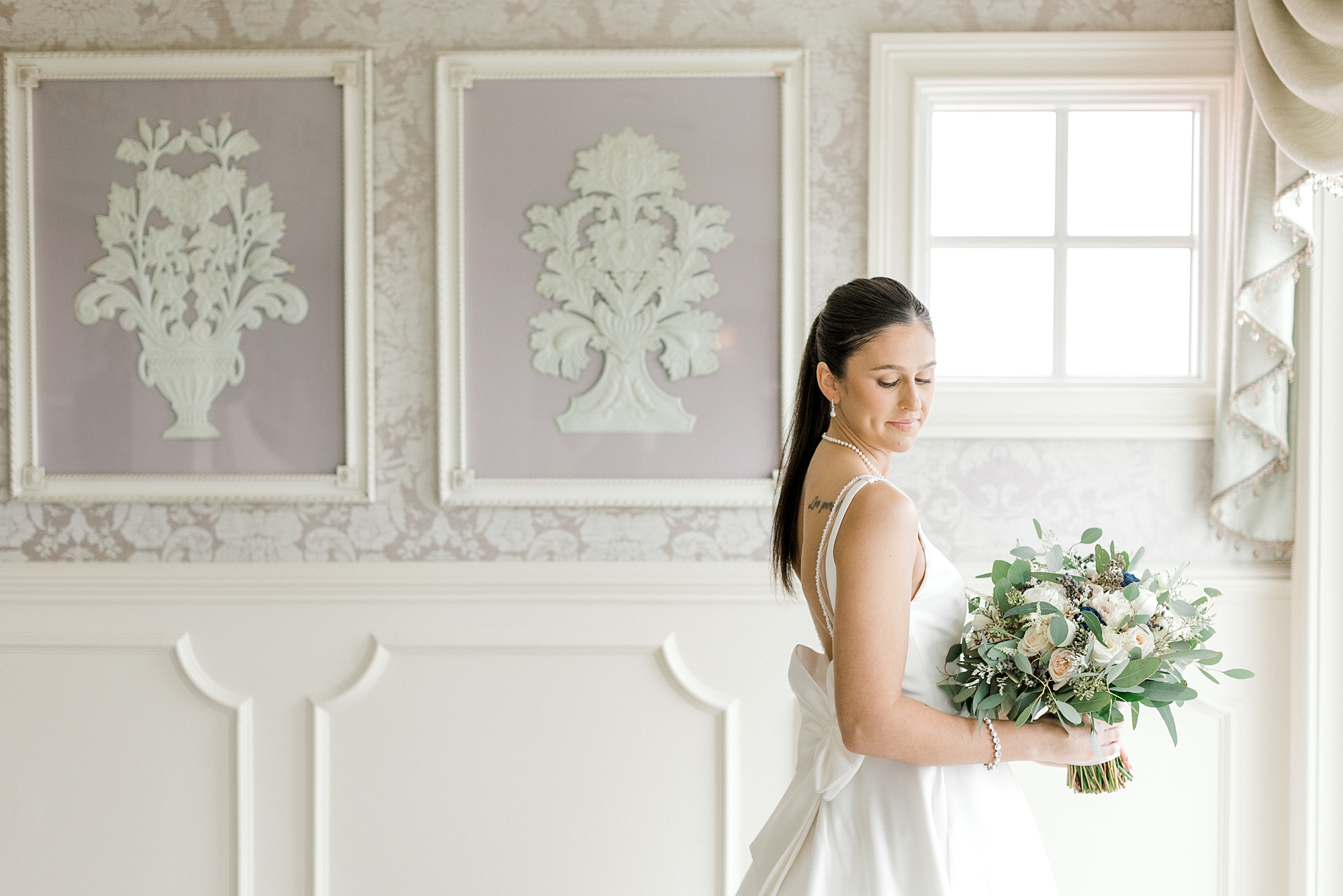 bride in wedding dress with bow on the back looks over shoulder holding bouquet of white and green flowers