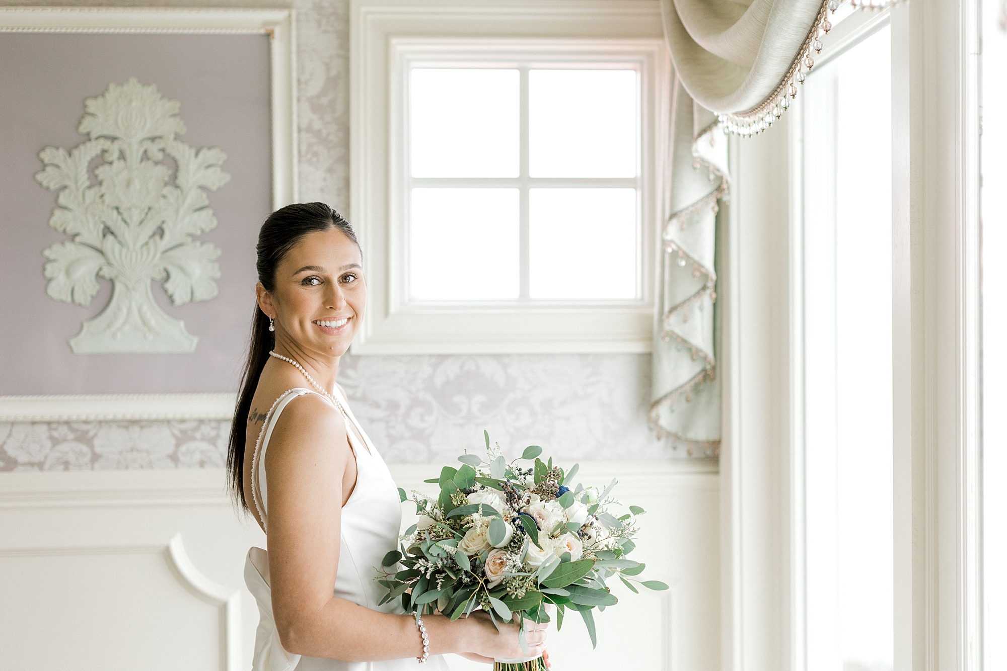 bride smiles holding bouquet of white flowers by window in suite at Mallard Island Yacht Club