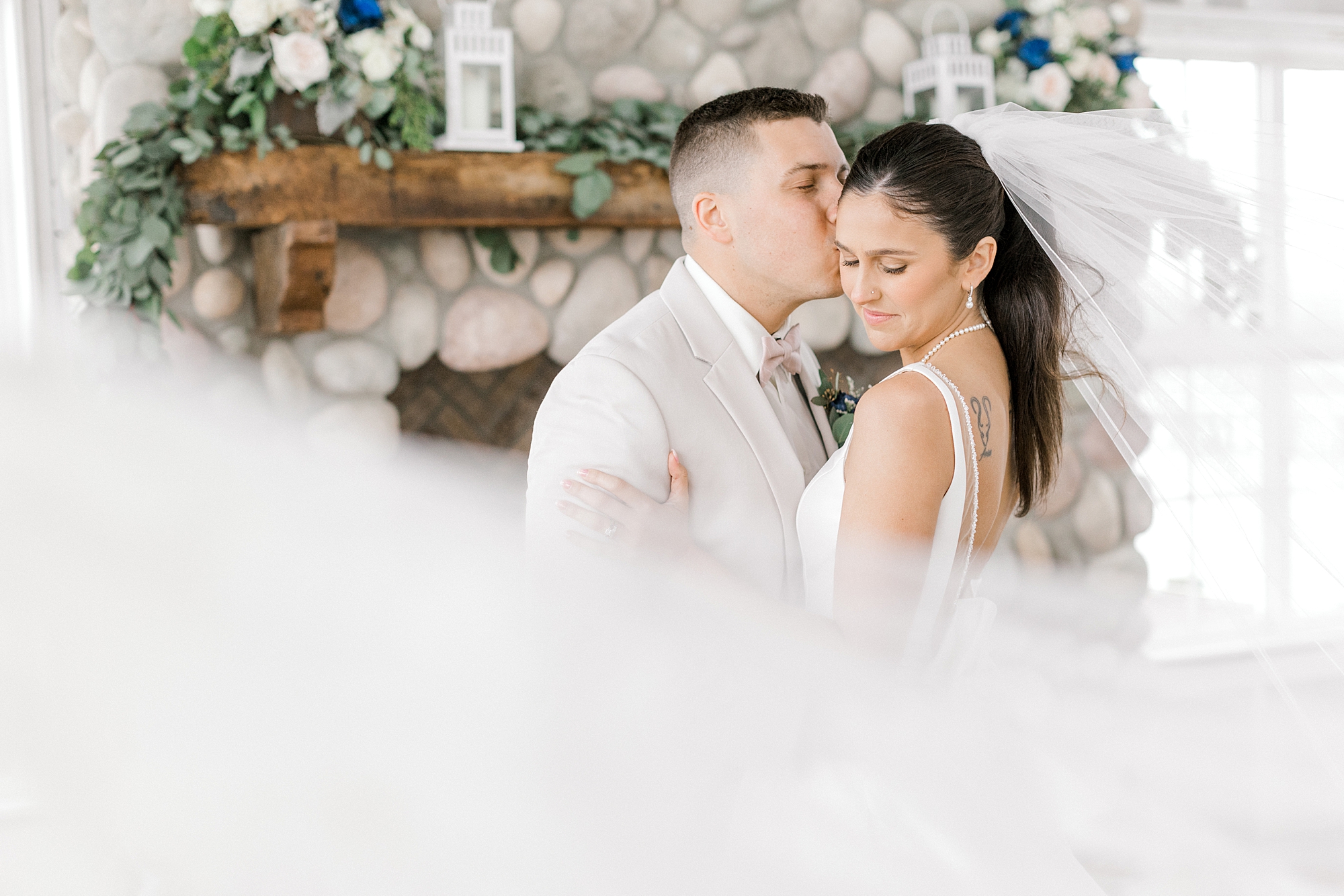 groom kisses bride's forehead with veil wrapped around them in front of stone fireplace at Mallard Island Yacht Club