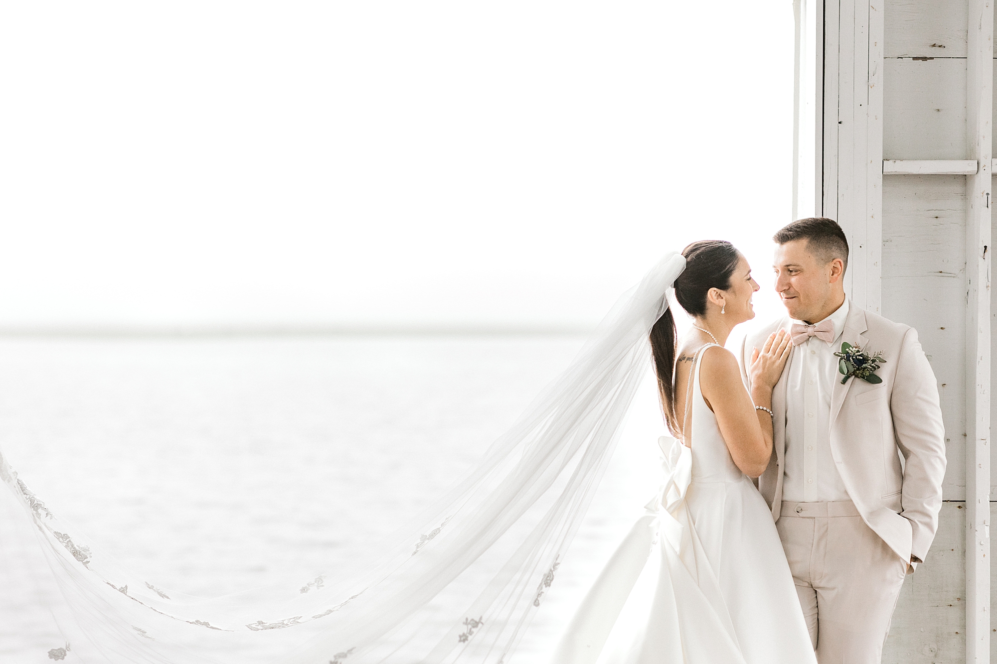bride looks up at groom while veil floats behind them by window at Mallard Island Yacht Club
