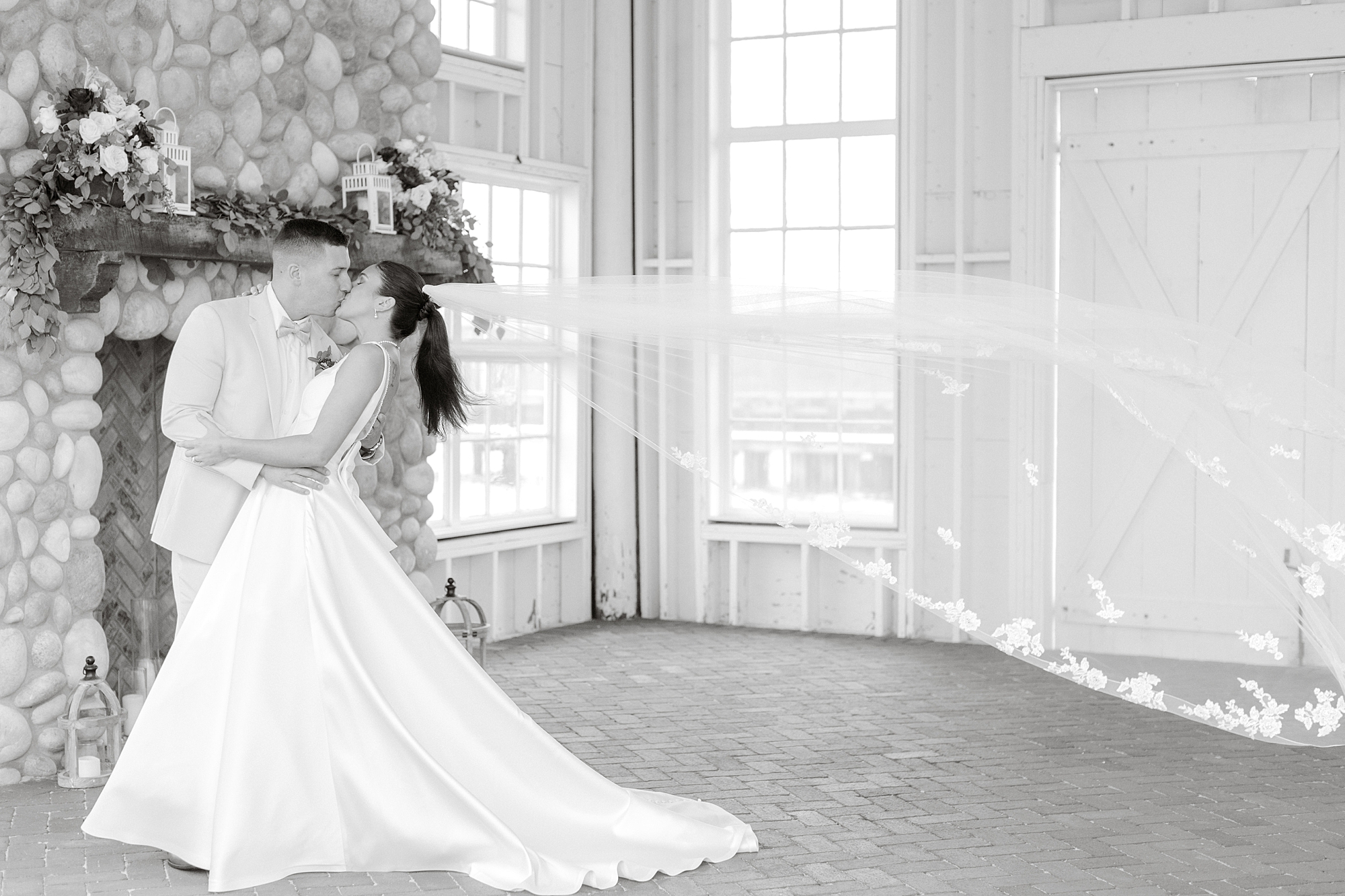 newlyweds kiss inside Mallard Island Yacht Club by stone fireplace while bride's veil floats behind them