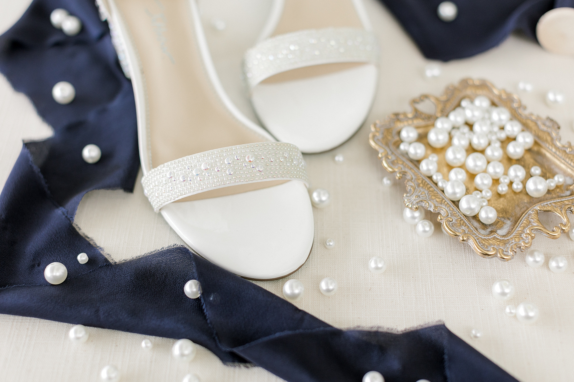 bride's pearl jewelry and white shoes lay near navy ribbon
