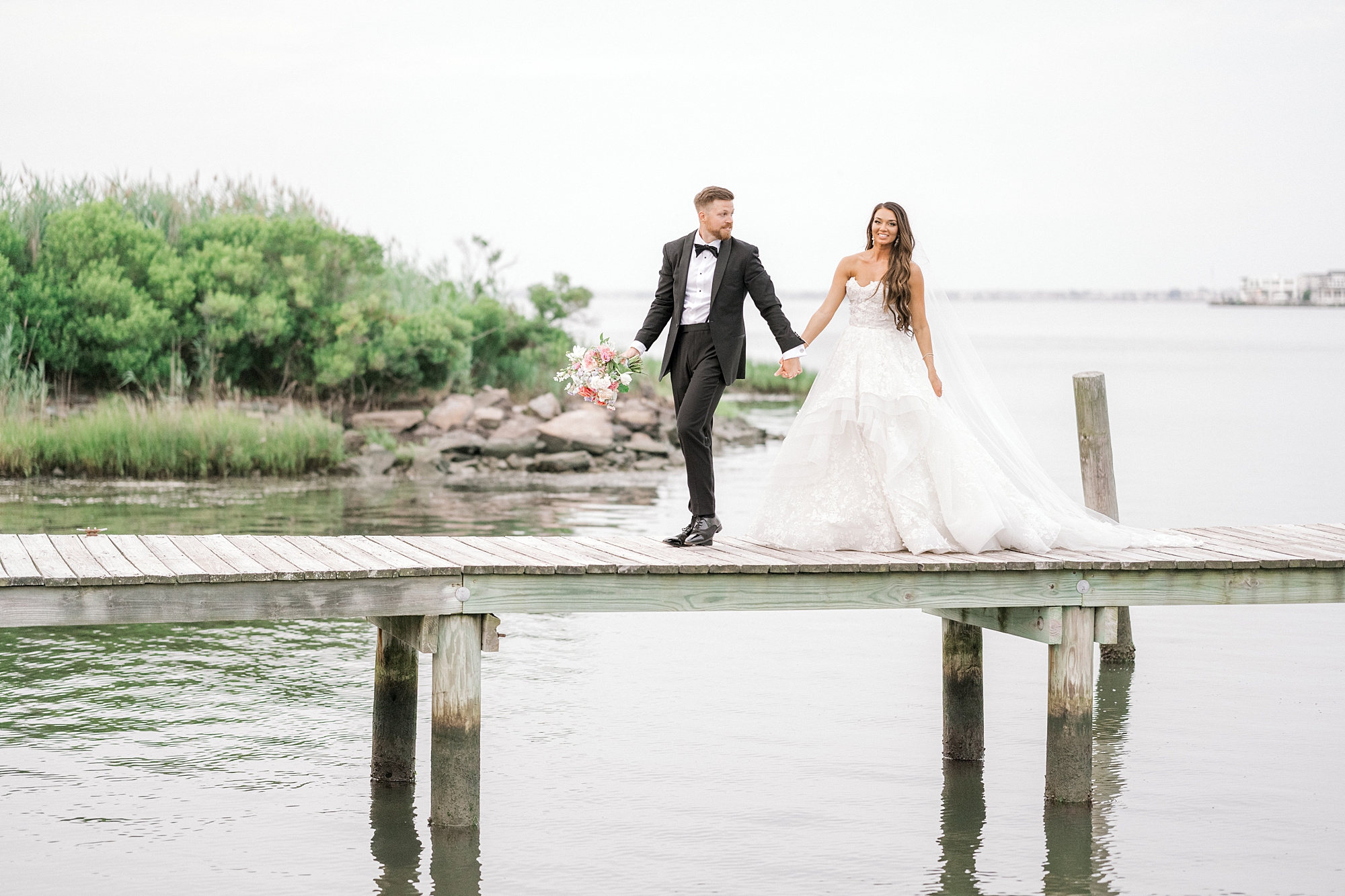 groom in tux leads bride in strapless wedding gown across wooden dock at Bonnet Island Estate on Long Beach Island