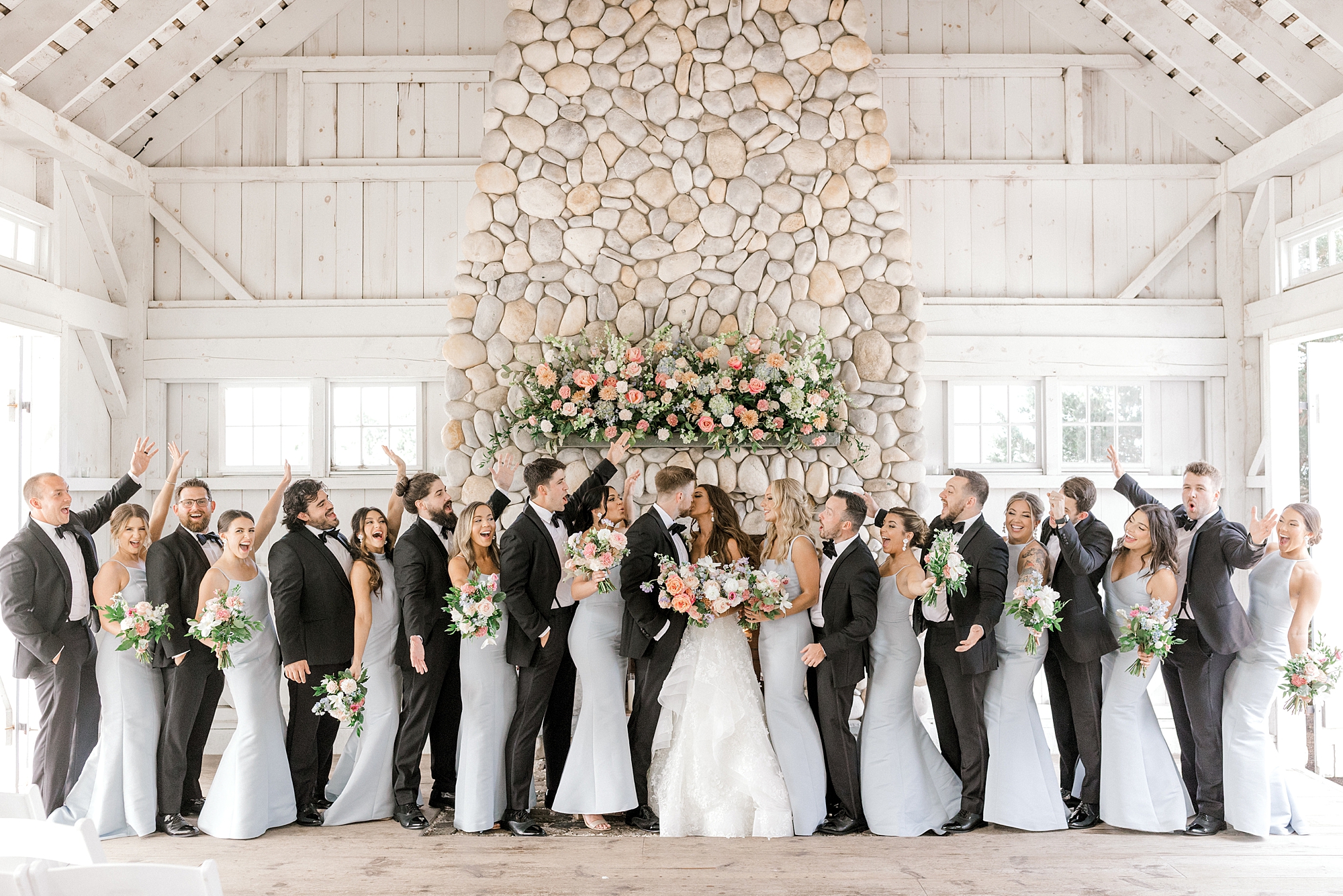 bride and groom kiss while wedding party in black suits and light blue gowns cheer in front of stone fireplace at Bonnet Island Estate