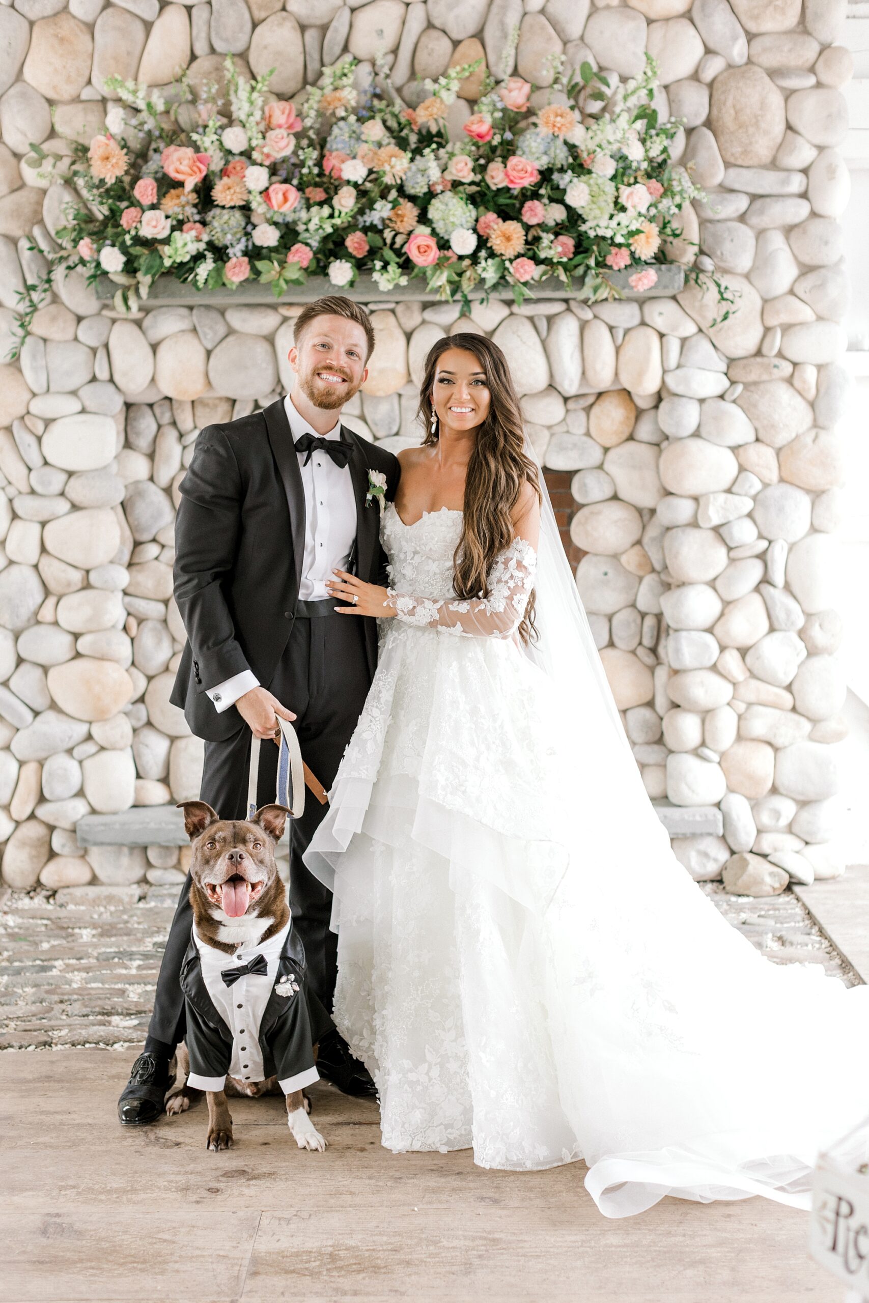 newlyweds pose by stone fireplace with dog sitting in front of them