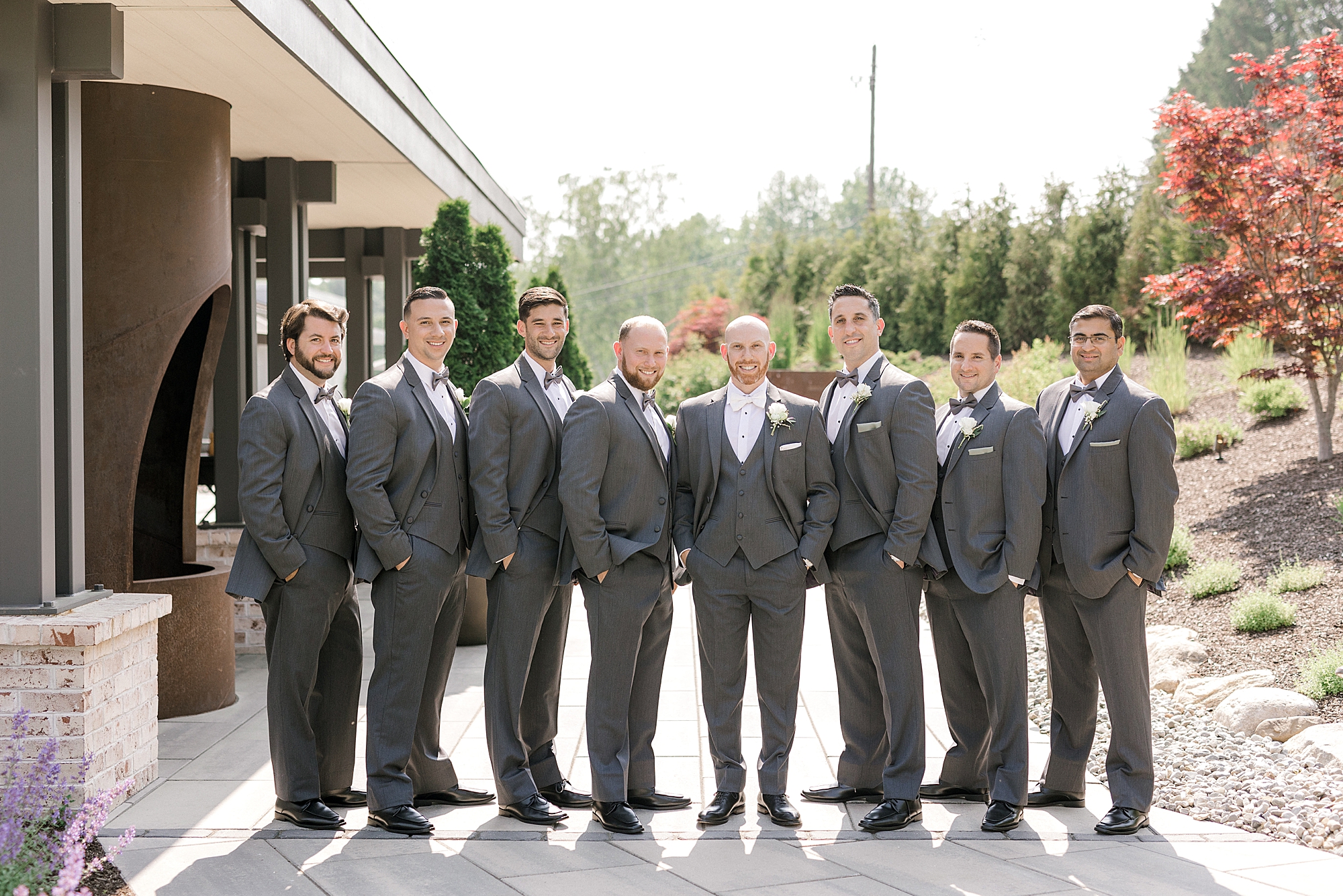 groom stands with groomsmen in grey suits on sidewalk at the Refinery at Perona Farms