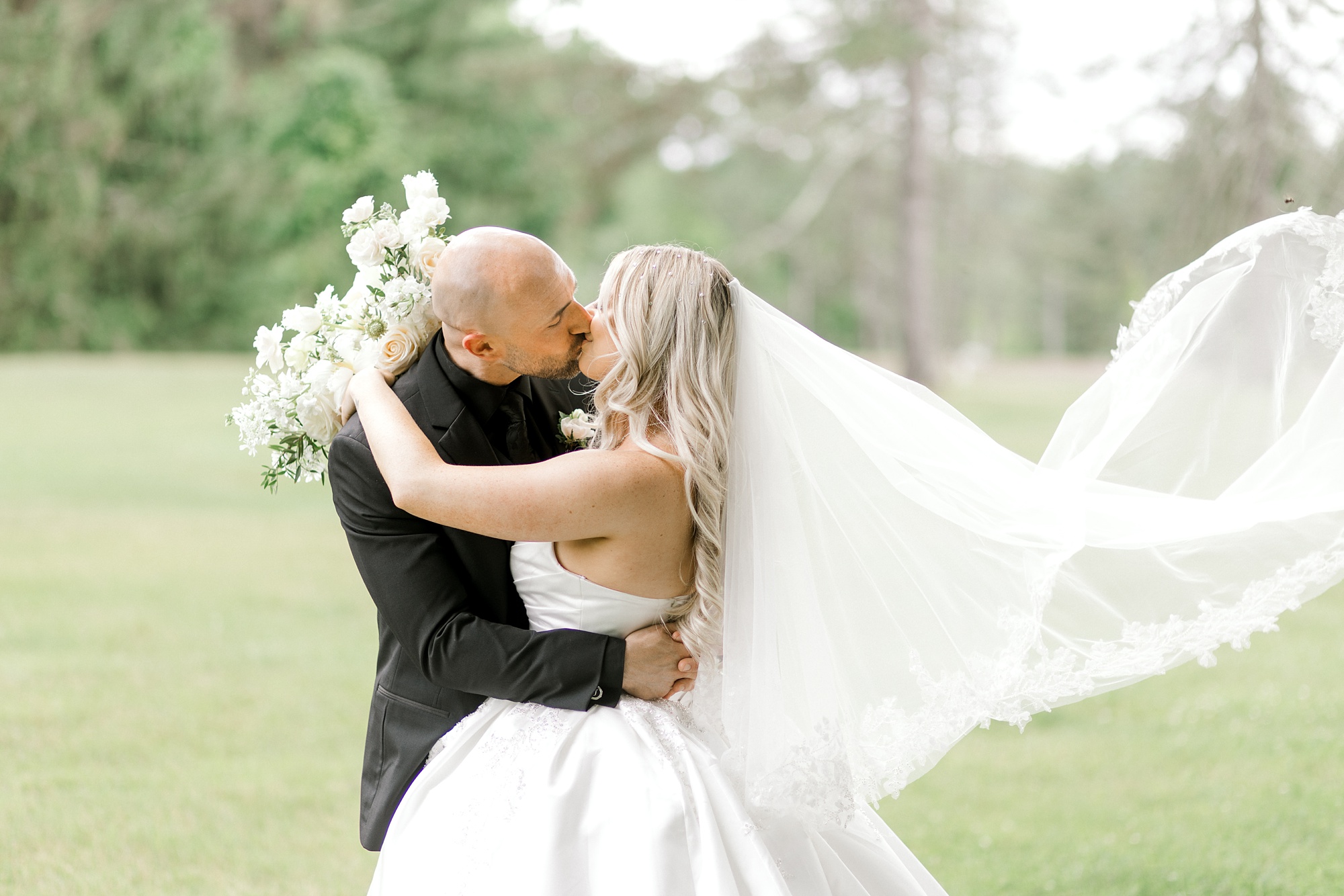 newlyweds kiss on lawn at the Refinery at Perona Farms with bride's veil floating