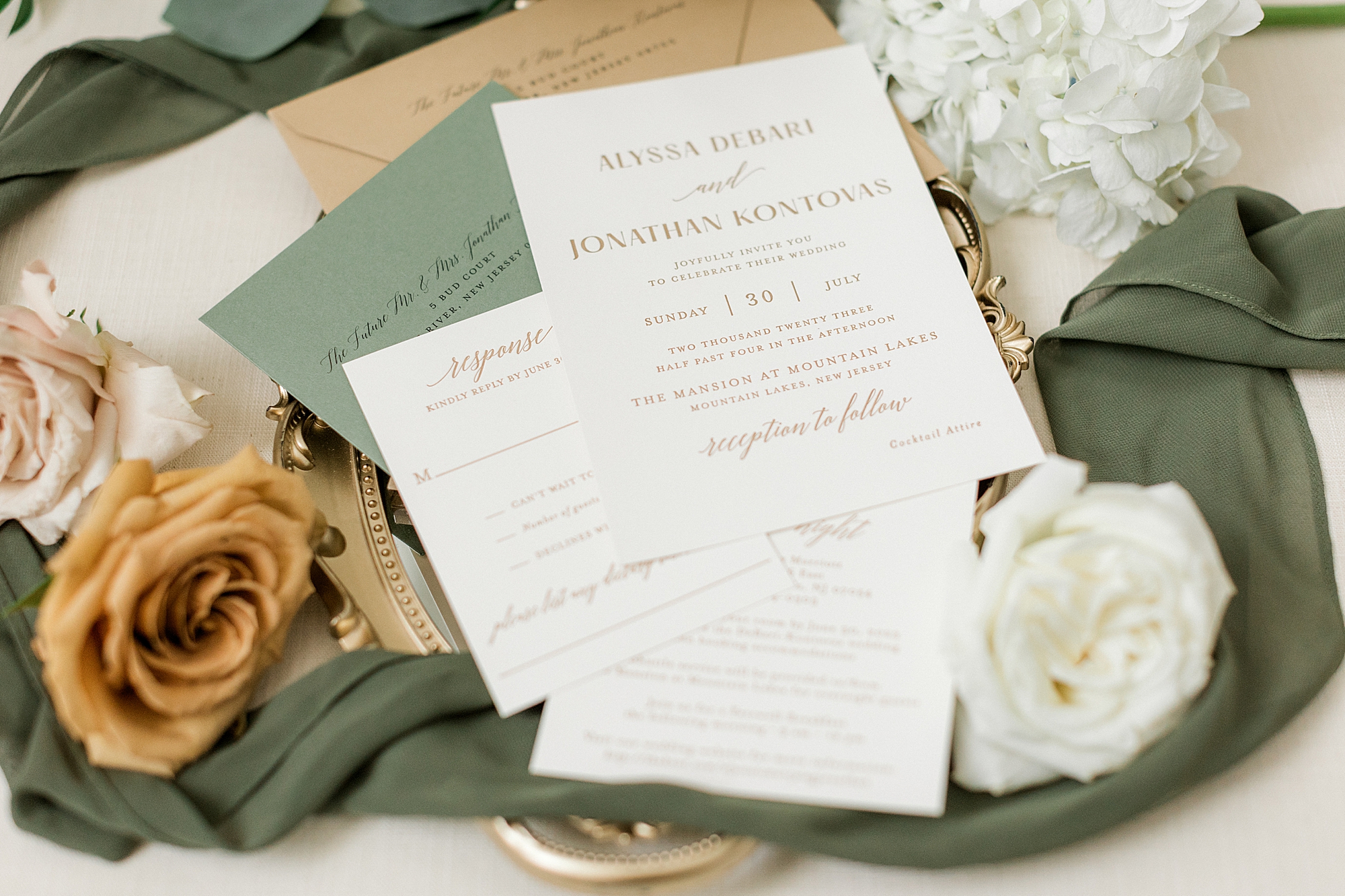 tan and forest green invitation suite lays with white and pale pink roses