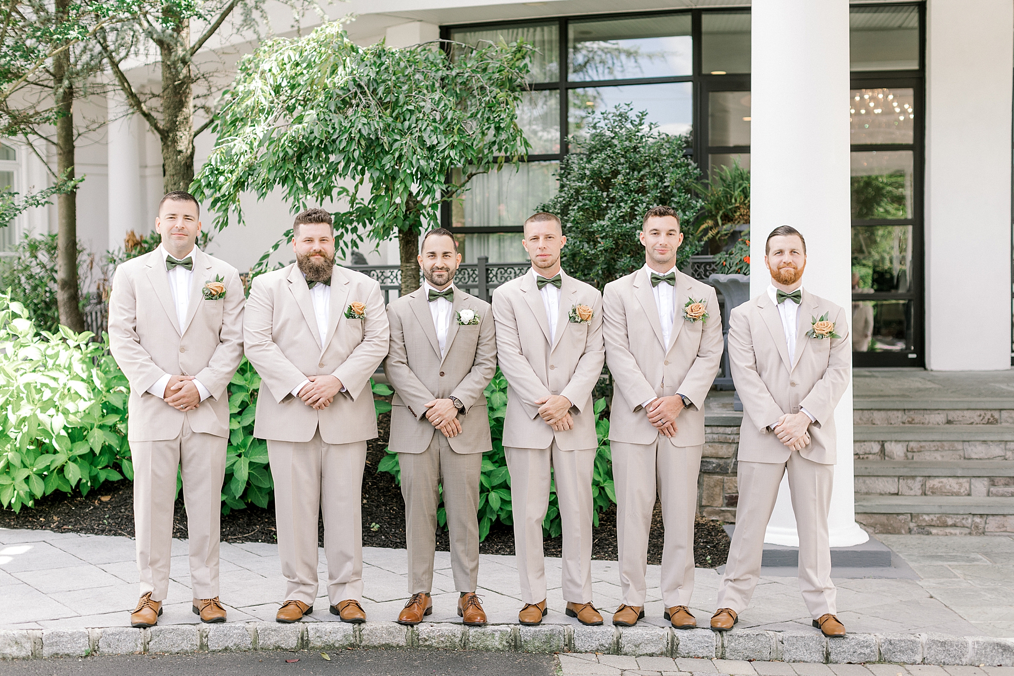 groom and groomsmen in tan suits stand with hands in front of them outside the Mansion at Mountain Lakes