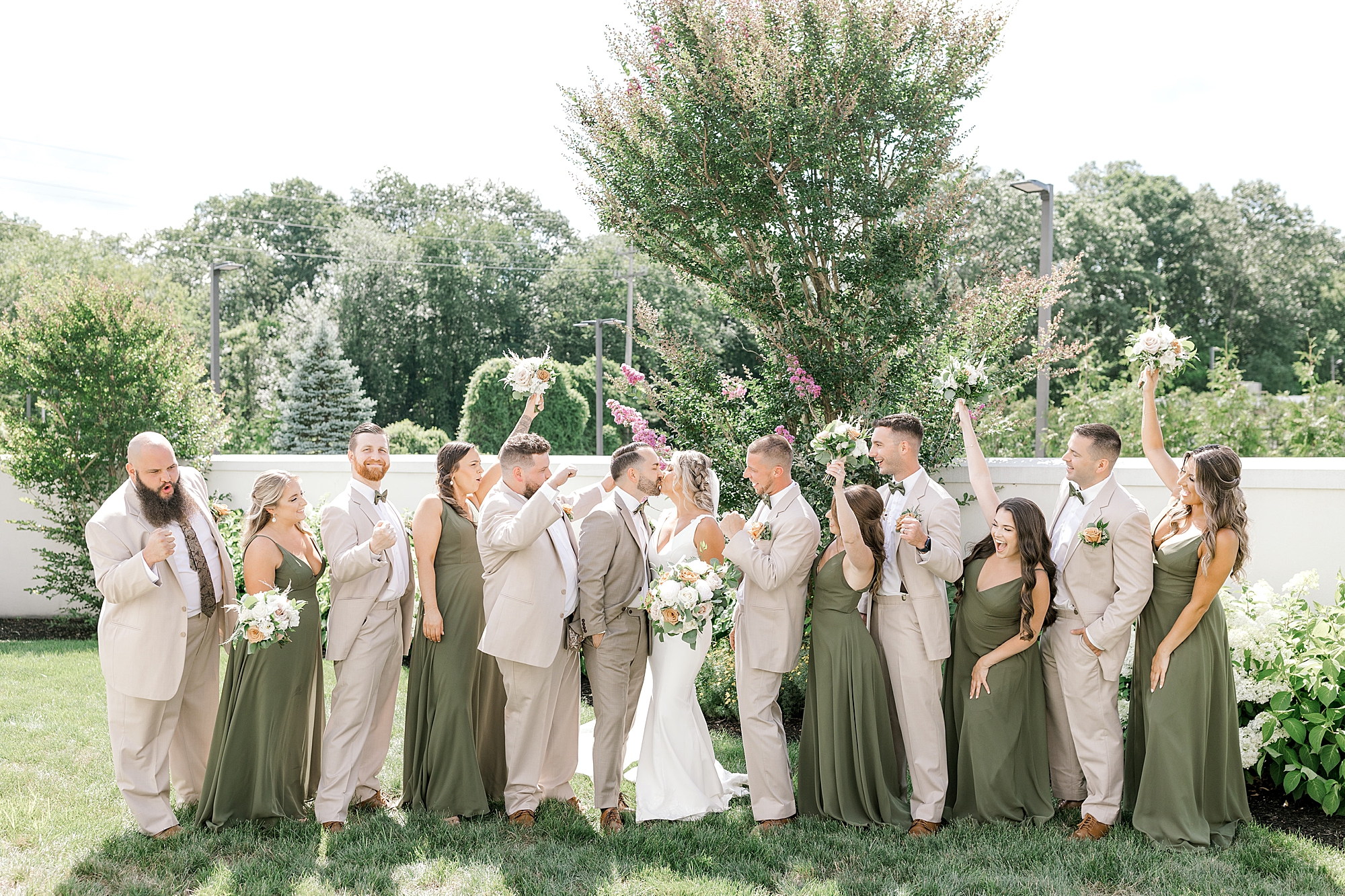 bridesmaids hold up bouquets cheering between groomsmen while bride and groom kiss during NJ wedding day