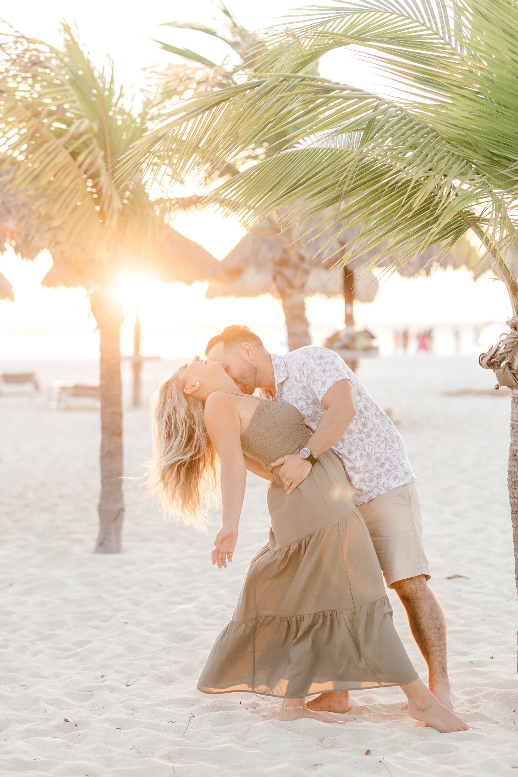 man kisses wife's throat on beach at sunset with trees through palm trees