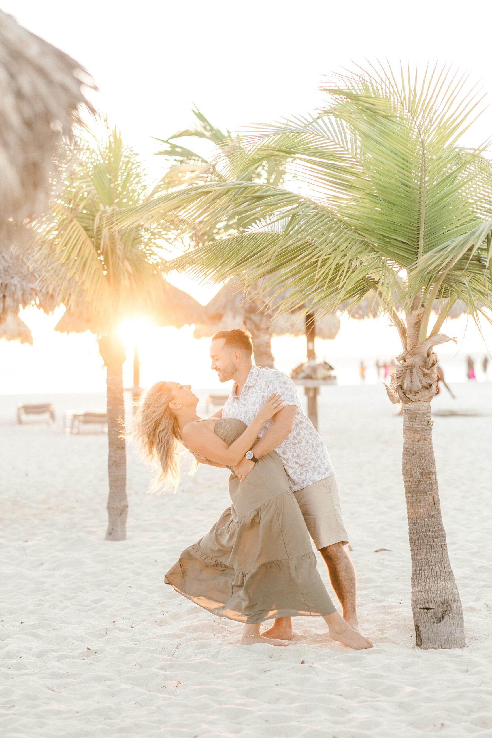 man dips wife on beach at sunset with sun coming through palm trees