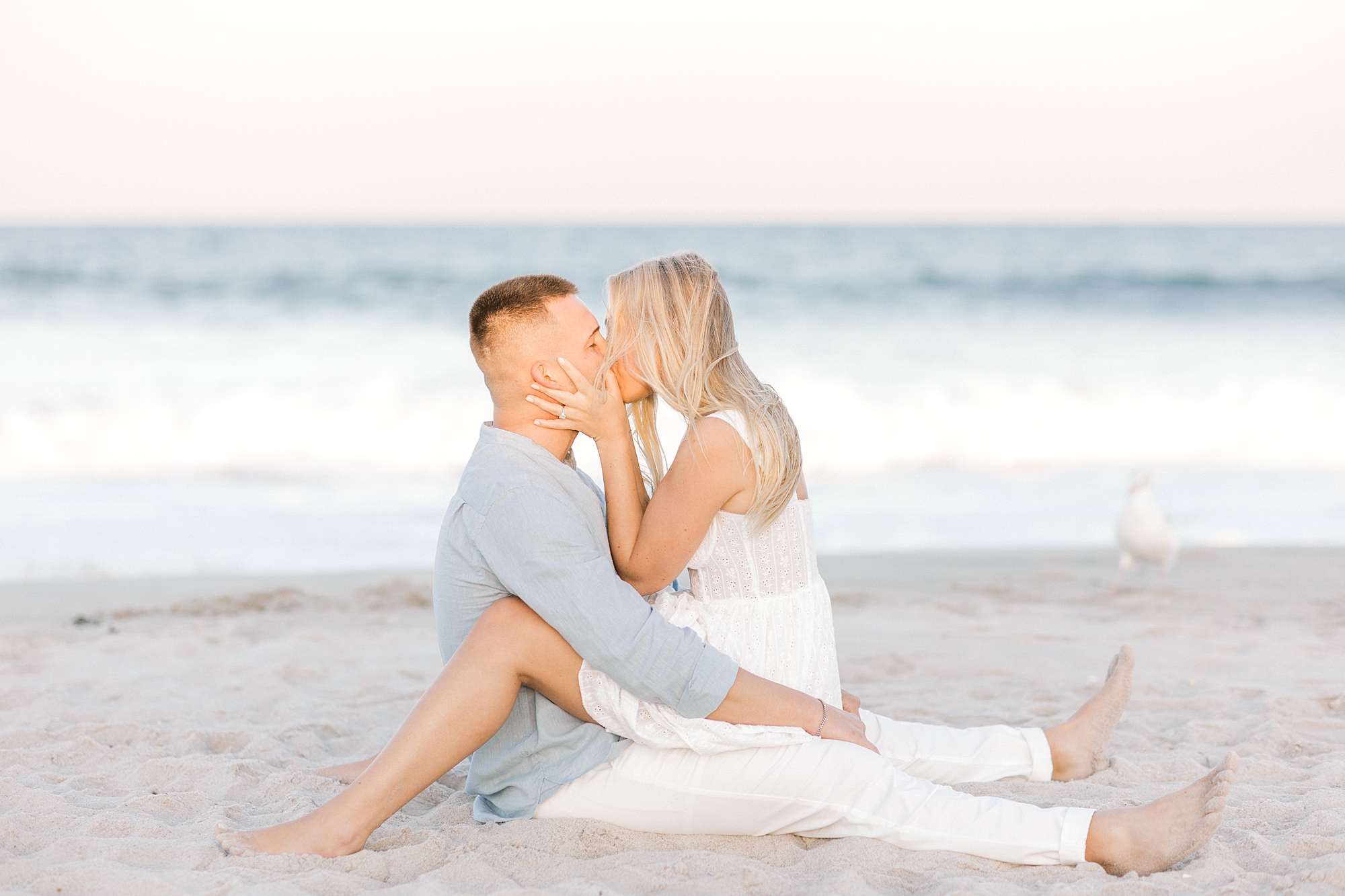 woman sits on man's lap in front of ocean during engagement session in Lavallette, NJ