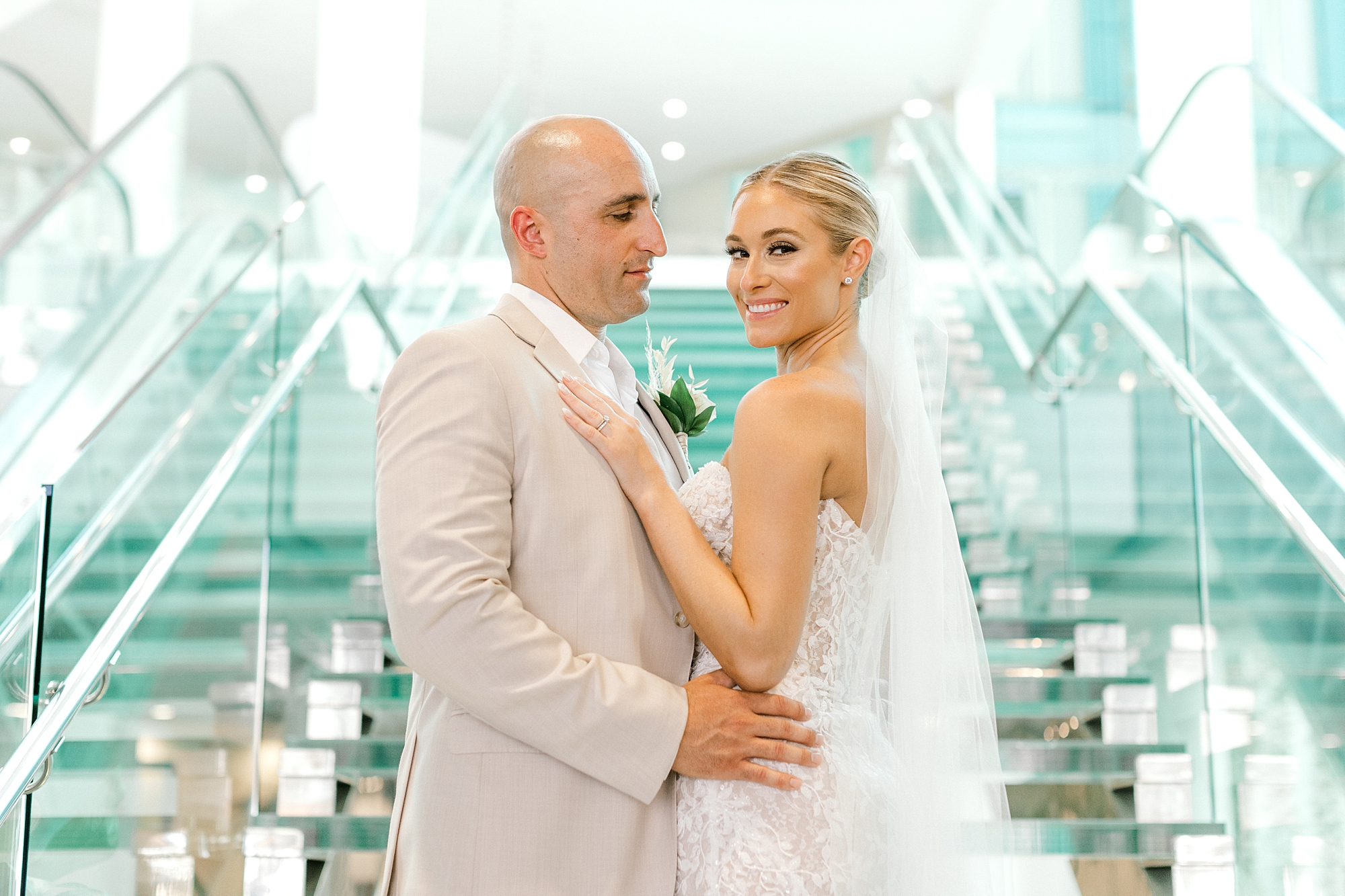groom in tan suit looks at bride on glass steps in hotel lobby in Aruba