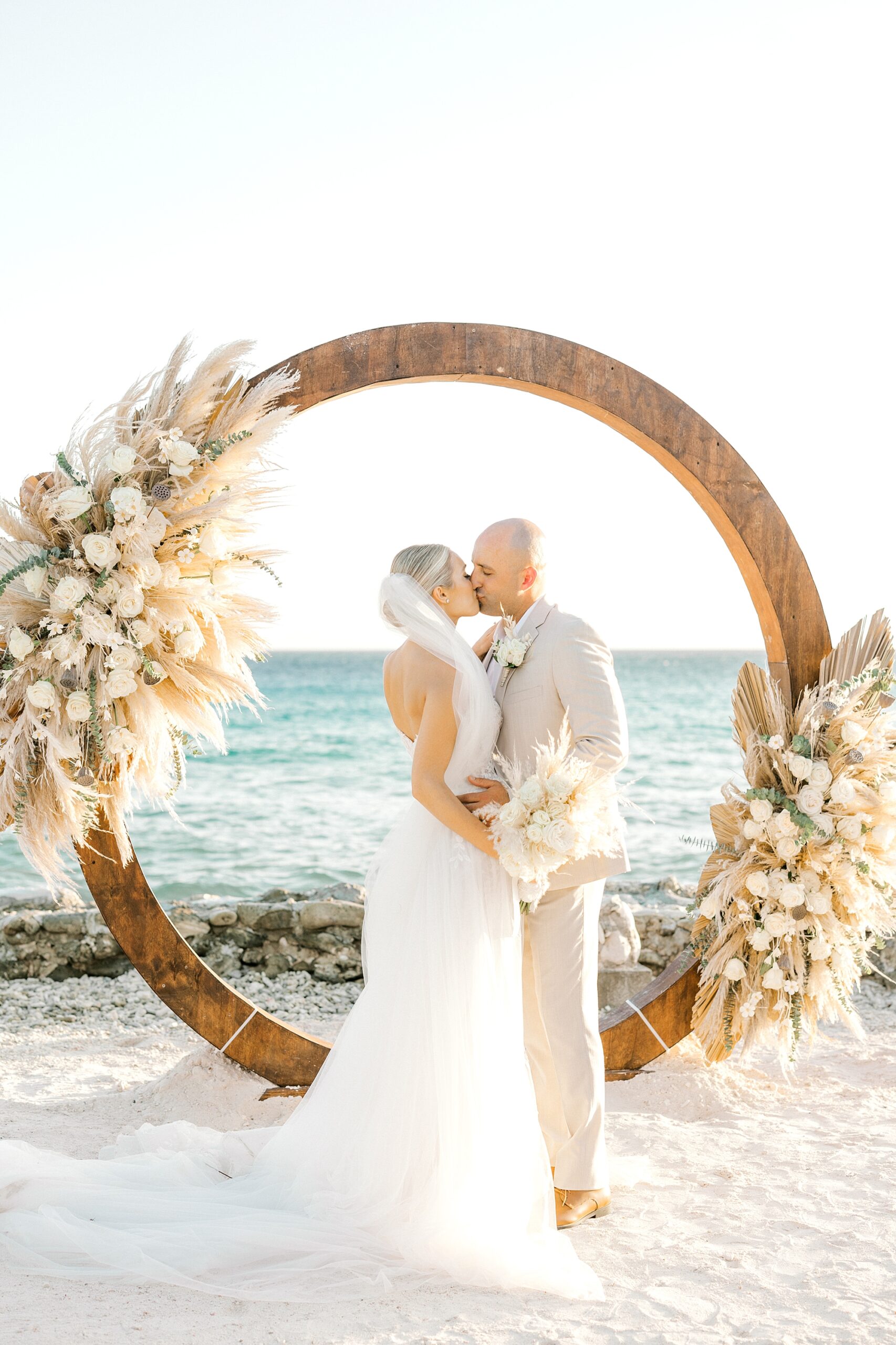 bride and groom kiss in front of wooden circle arbor with ivory and tan flowers
