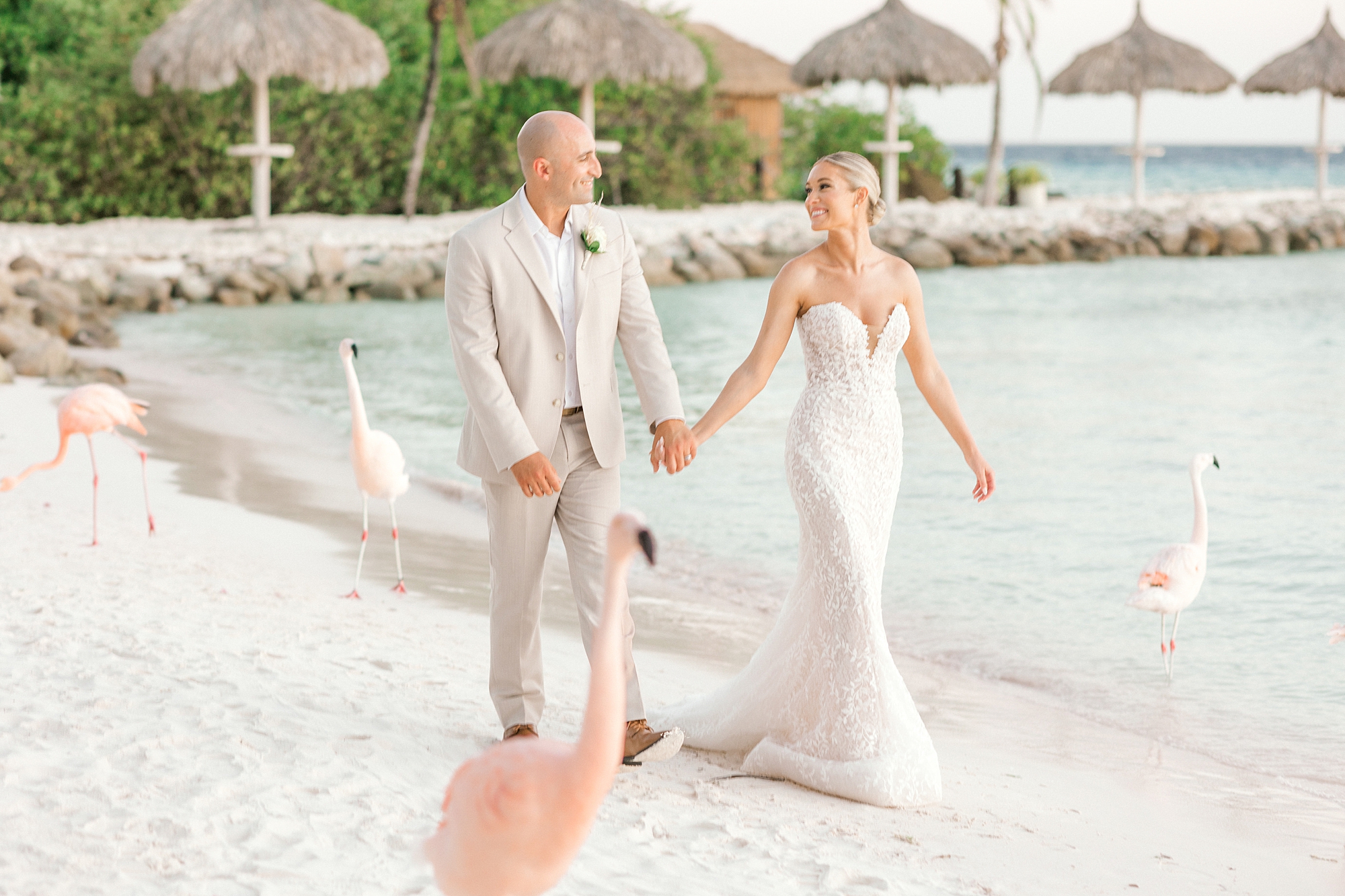 bride and groom hold hands walking on beach with flamingos