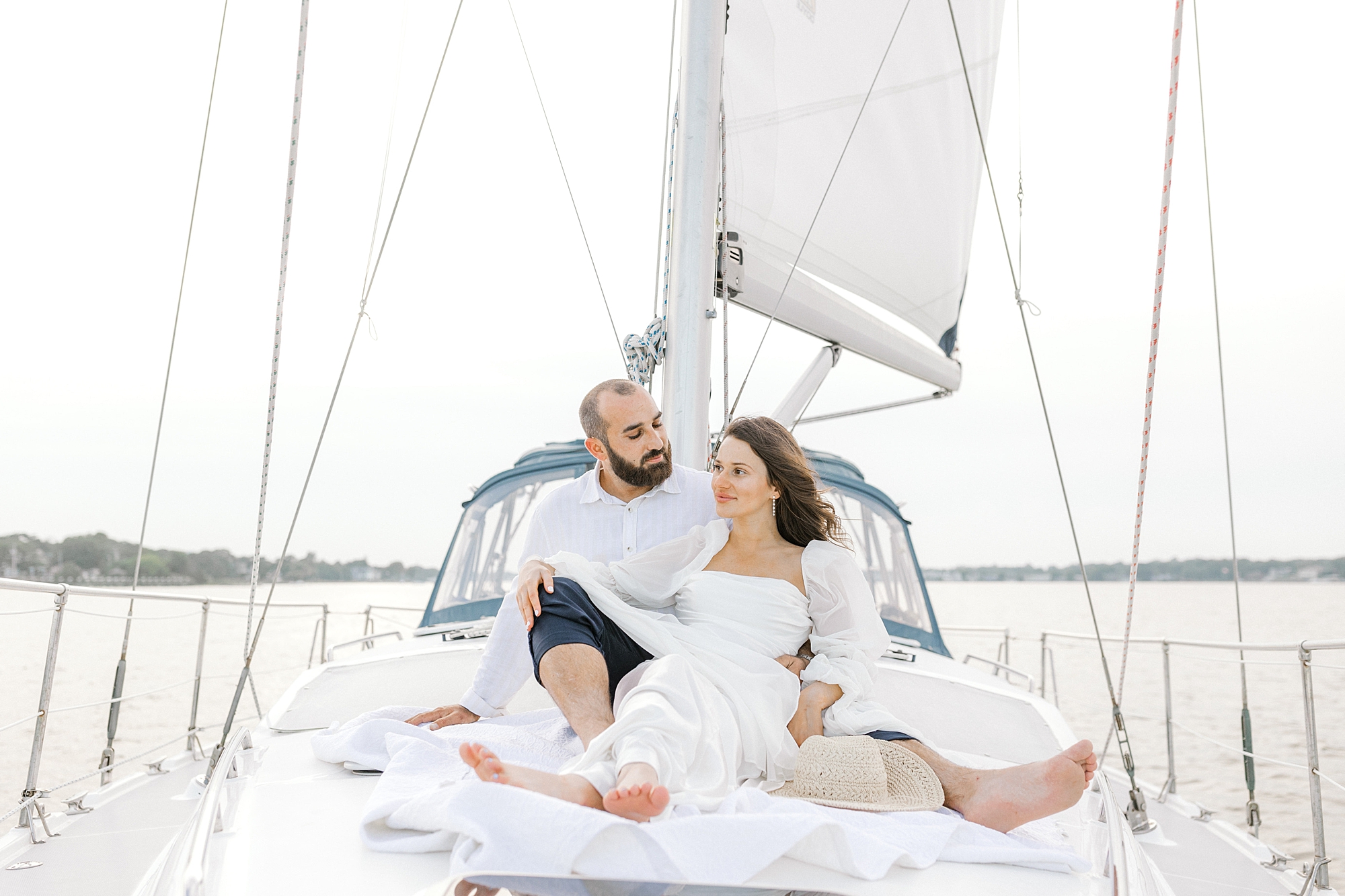 woman in white dress sits in front of man on front of sailboat during Toms River engagement session