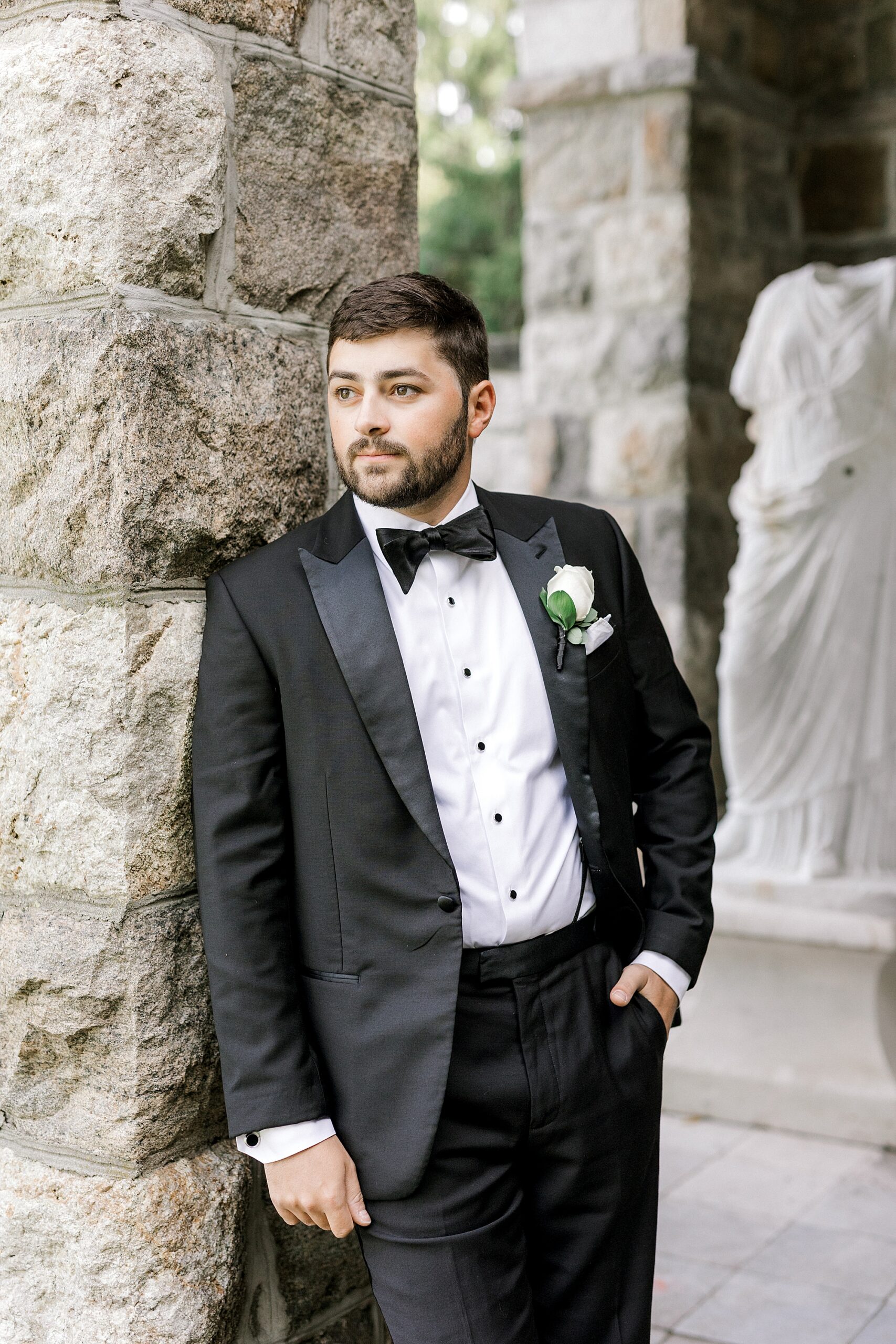 groom leans against stone wall in black tux in New Jersey