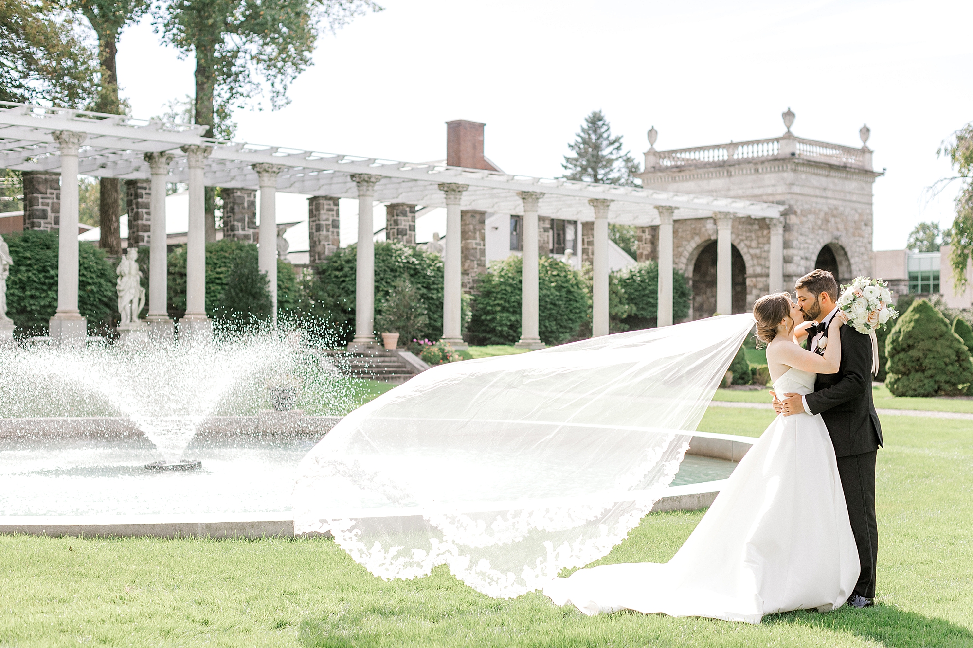 newlyweds kiss on lawn at the Farmhouse in New Jersey with bride's veil floating behind them