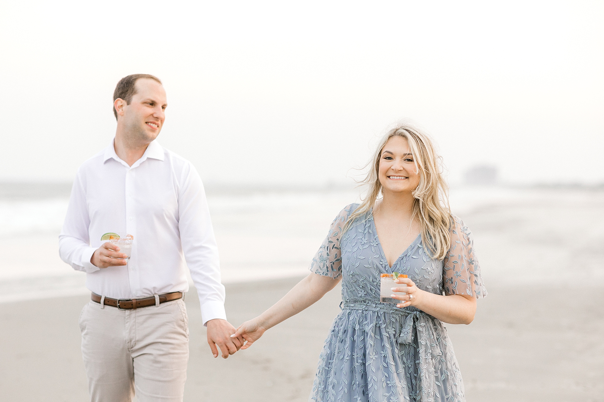 engaged couple holds hands and carries margaritas walking on beach during engagement session
