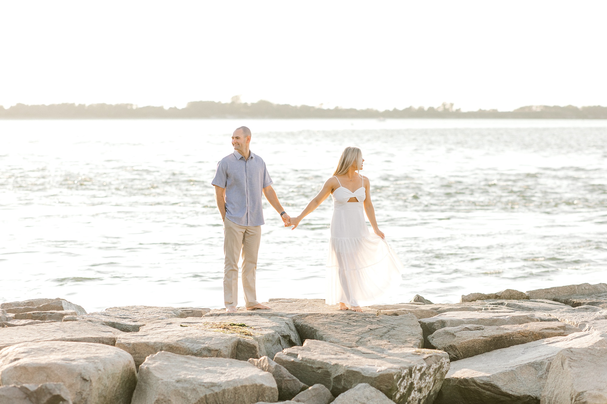 engaged couple holds hands standing on rocks in front of water on Long Beach Island