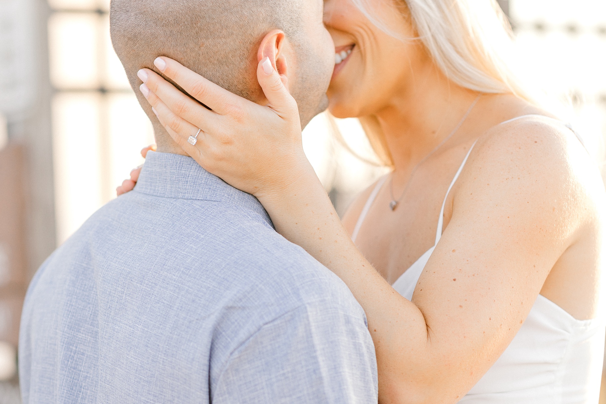 engaged couple lean to kiss with bride showing off engagement ring resting on groom's neck