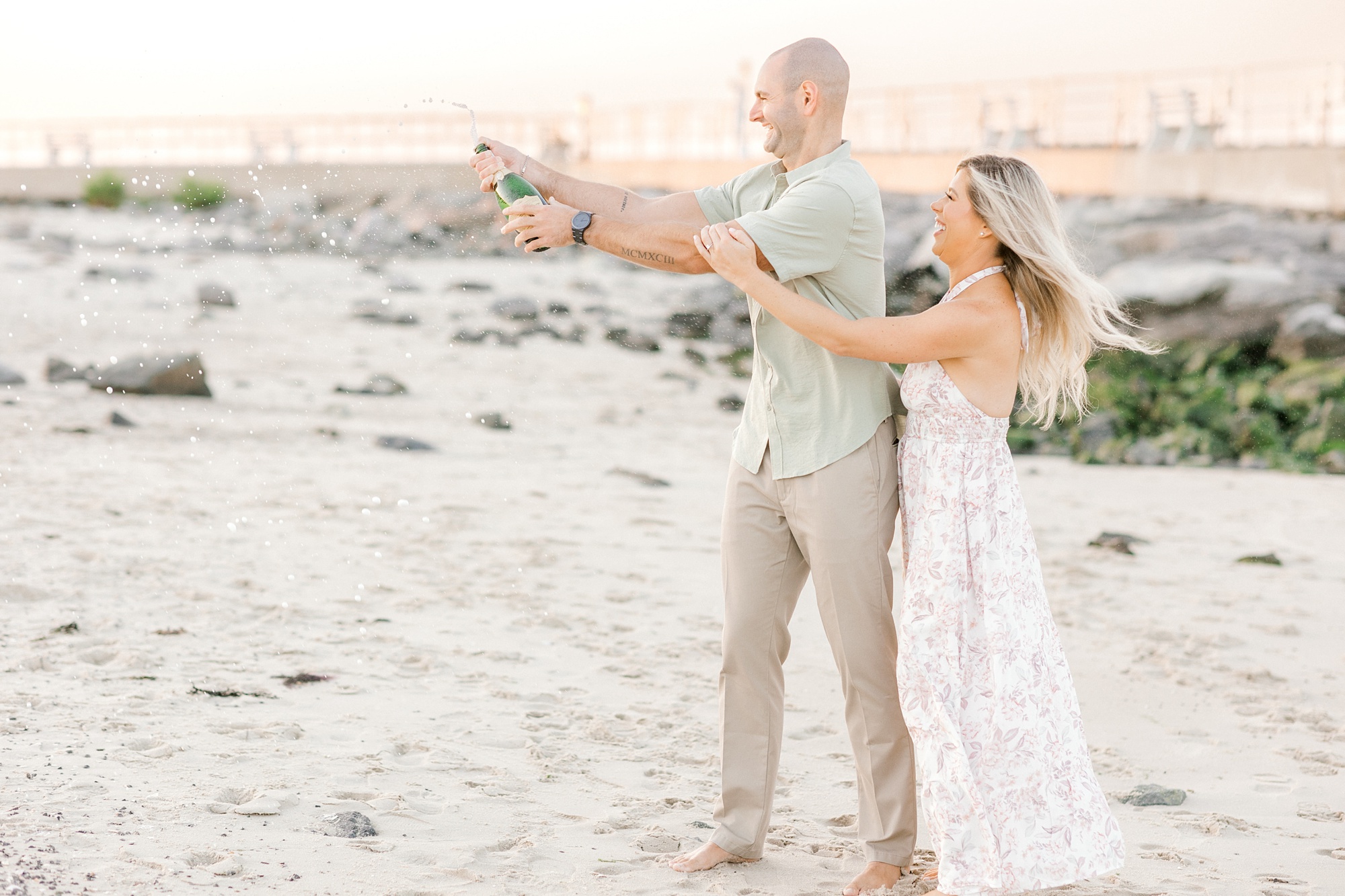 engaged couple pops champagne bottle at sunset near Barnegat Lighthouse