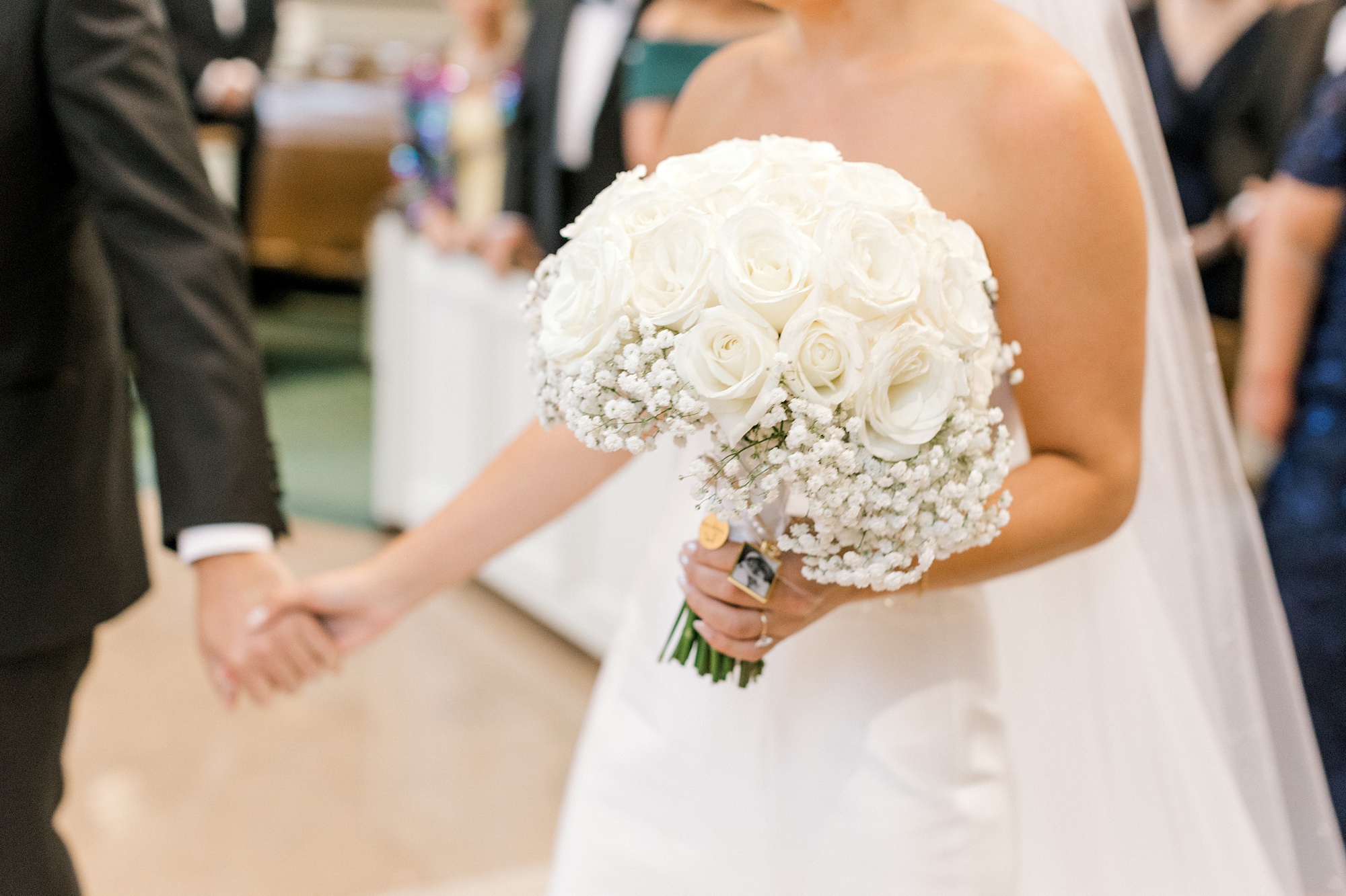 bride holds groom's hand and holds bouquet of white roses and baby's breath in the other