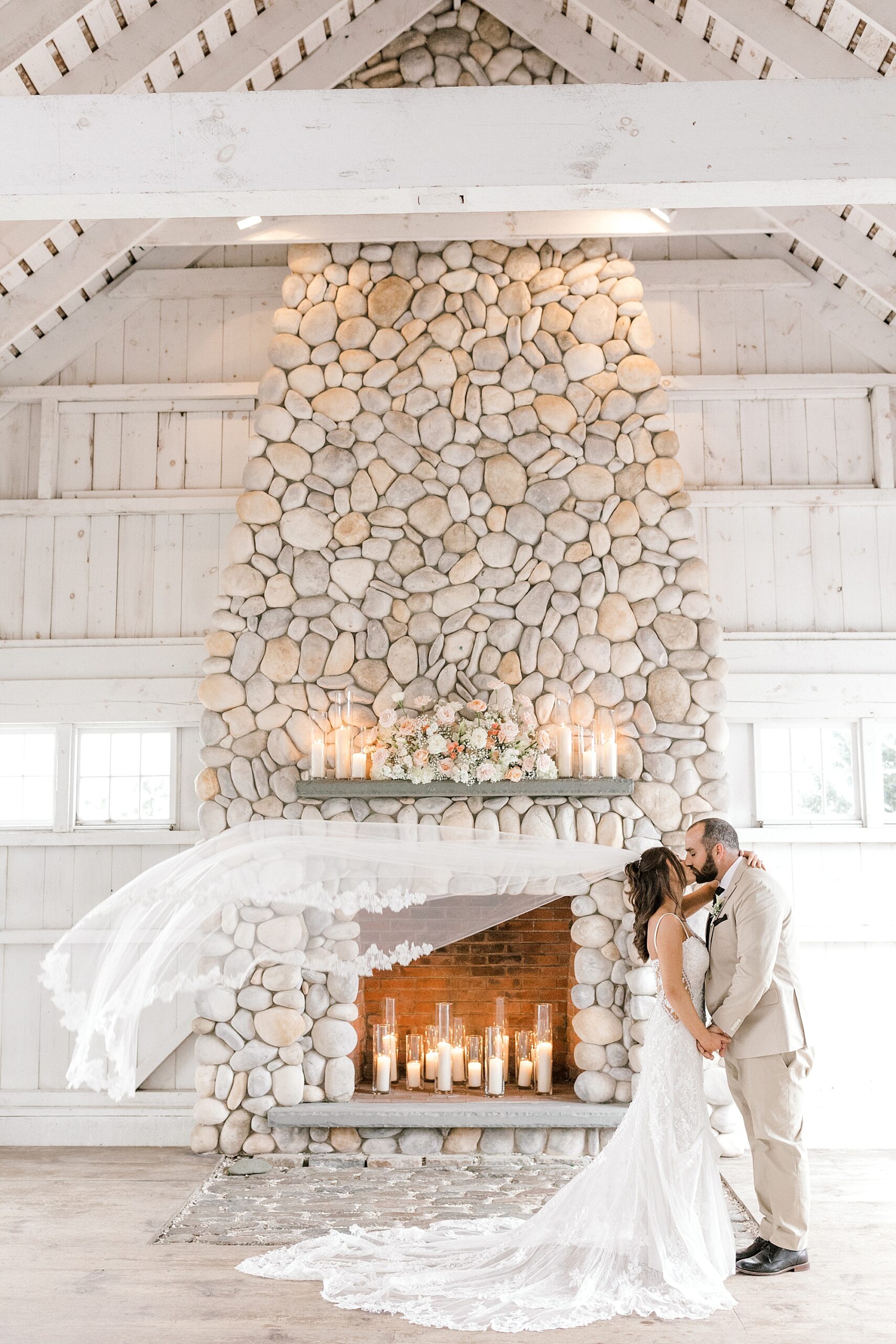 groom kisses bride in front of stone fireplace at Bonnet Island Estate with veil floating behind them