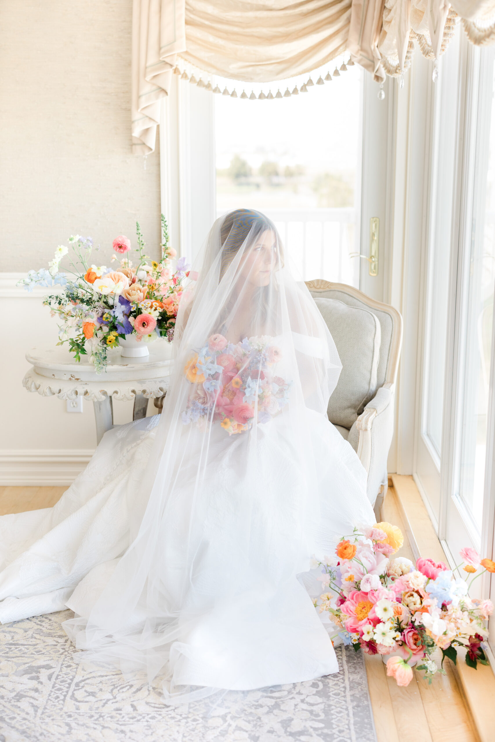 Bride holding bouquet sitting by window surrounded by colorful flowers and the veil over her face